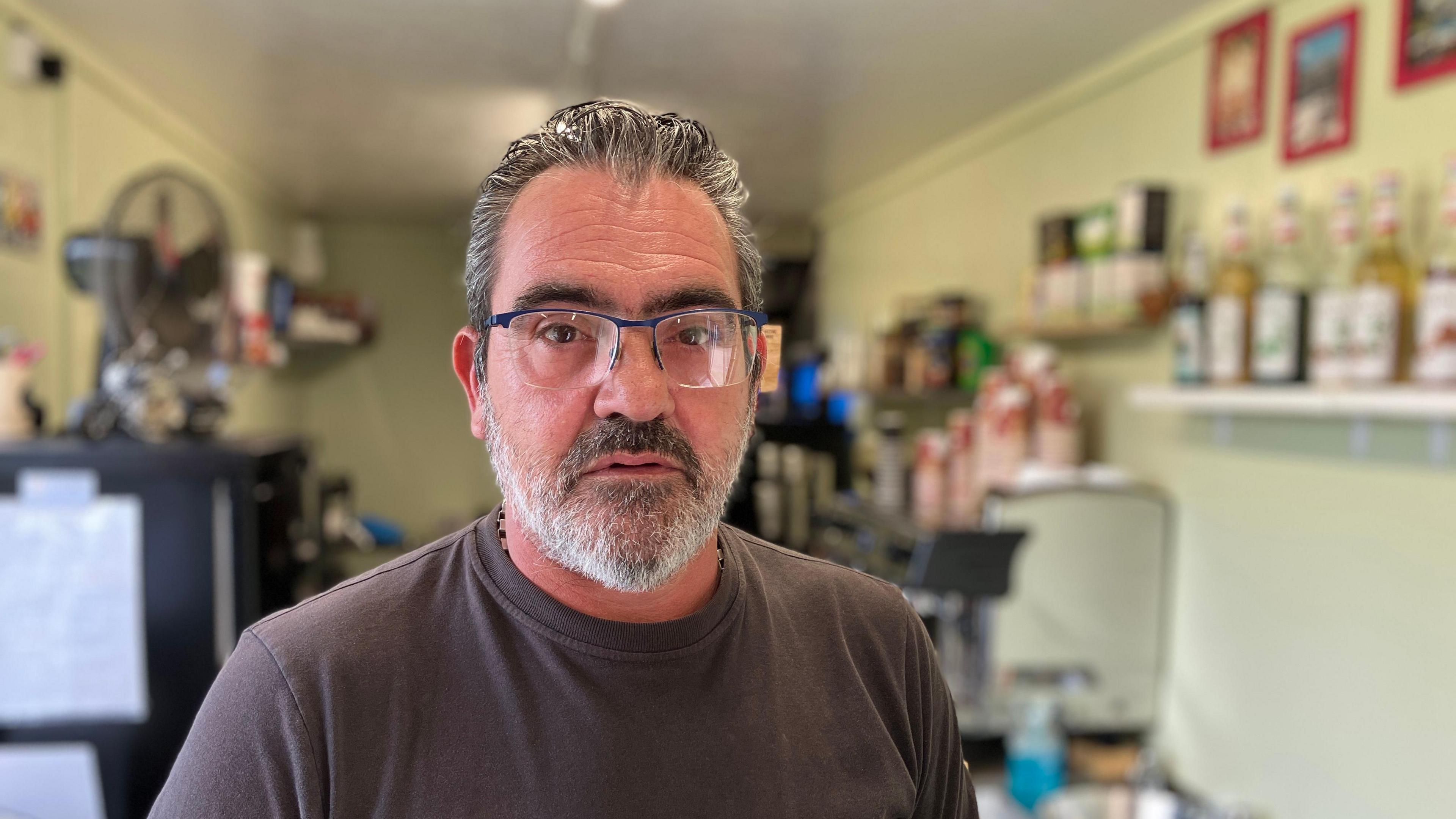 Bearded man in square glasses in a black top with a coffee shop in the background