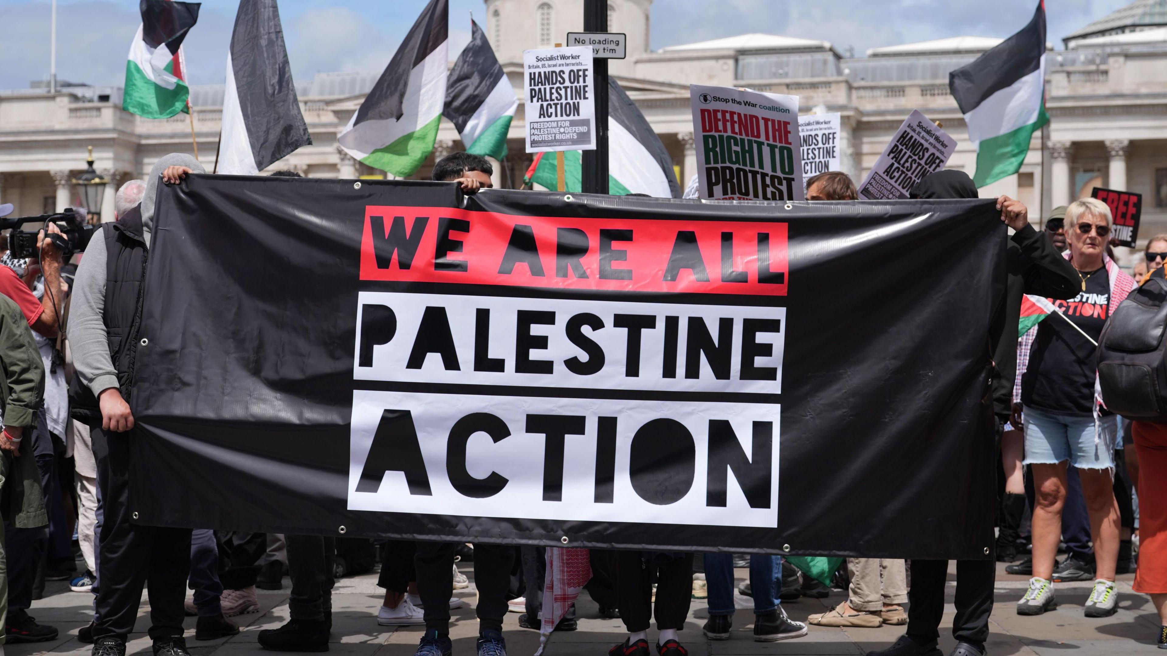A banner for Palestine Action being held up reading 'We Are All Palestine Action' - it is being held up at what appears to be a protest, with a number of Palestine flags visible in the background 
