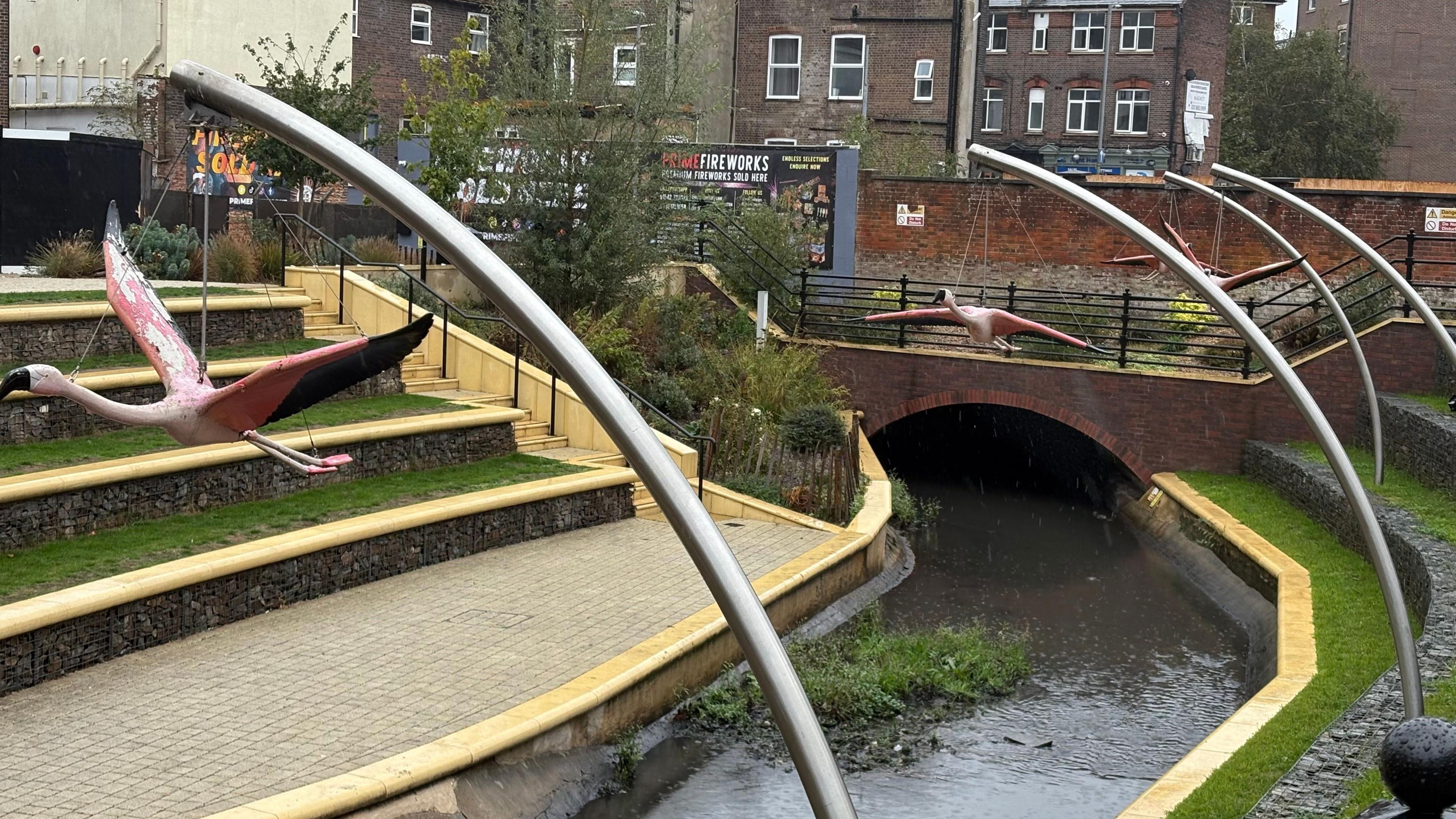 Brick-built bridge with a river flowing through. There are stepped seating areas on either side, with grass on each level. In the foreground, there are metal poles bearing models of large birds in flight. A three-storey brick building with small windows is visible in the background.