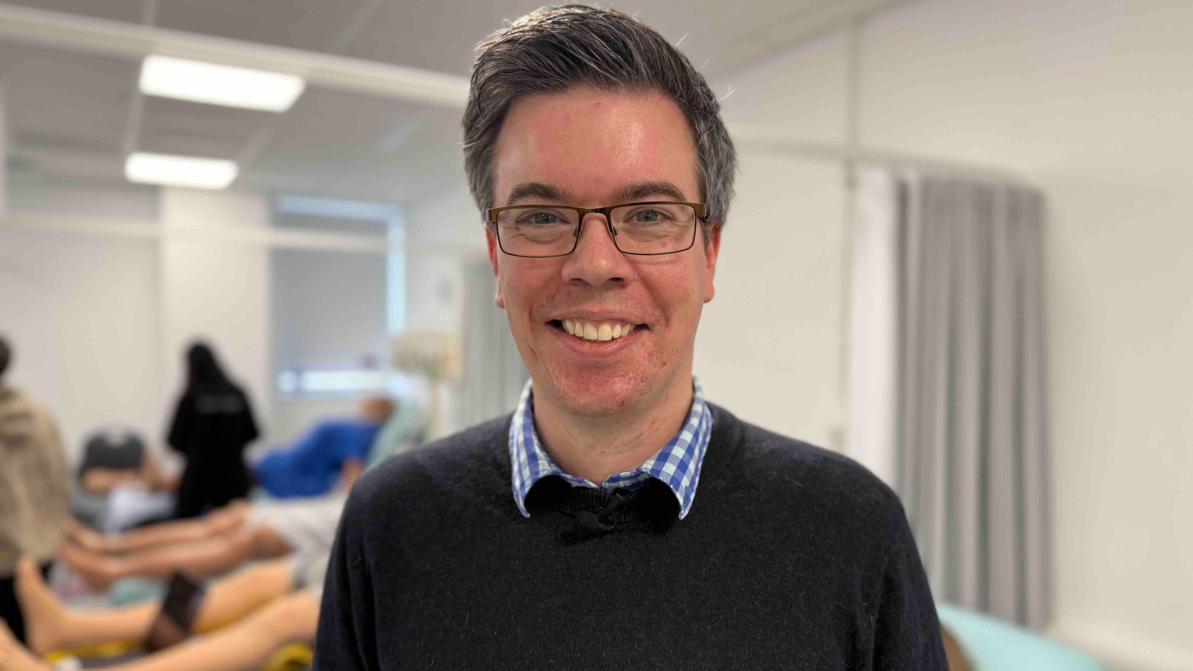 A man with short black hair, greying at the sides, smiles at the camera. He is wearing a black jumper over a blue chequered shirt and black-framed glasses.
The background is out of focus, but mannequins can be seen lying on hospital beds.