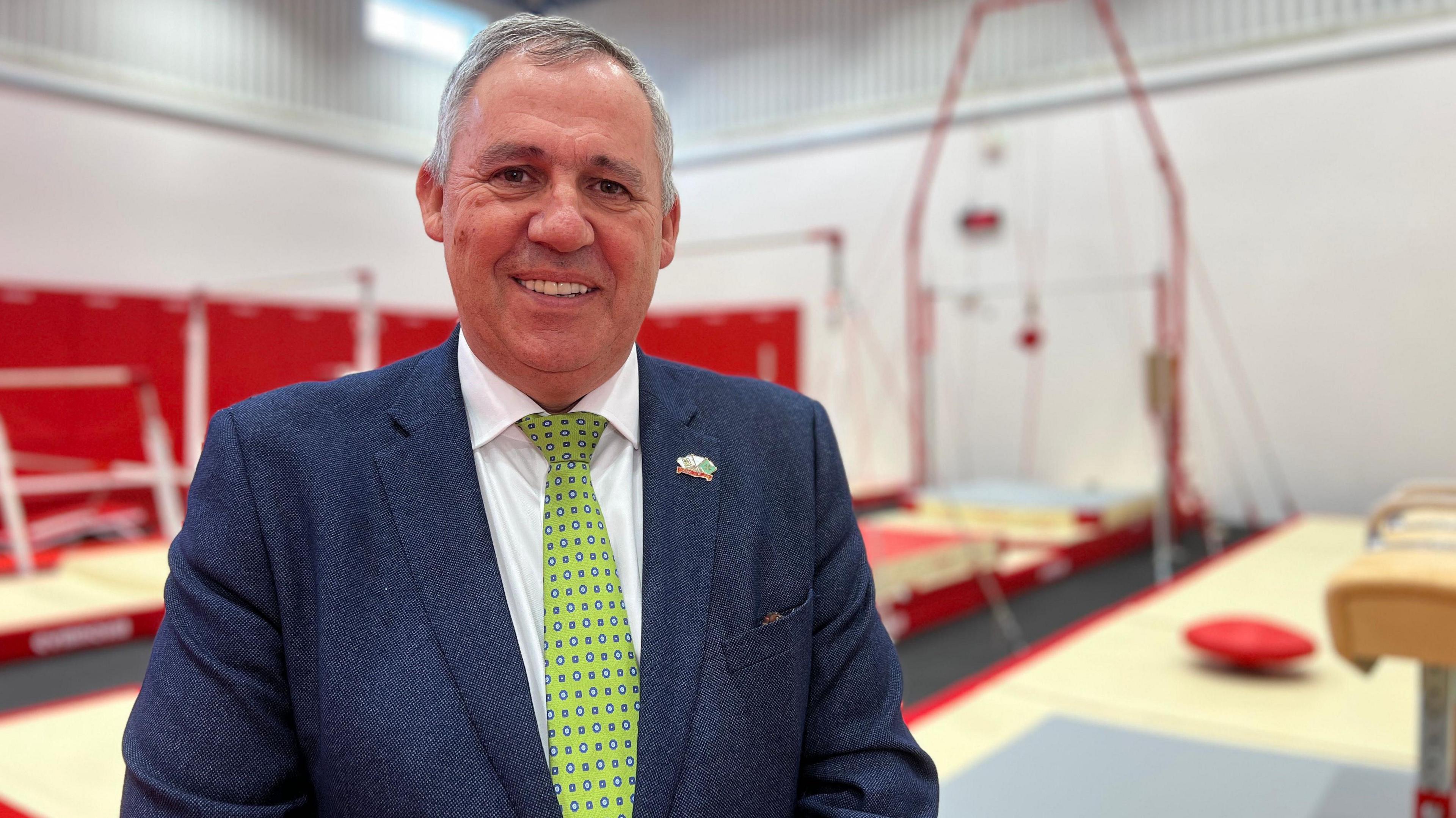 A man in a blue suit with a green tie stands in front of gymnasium equipment in a sports hall. He is smiling.