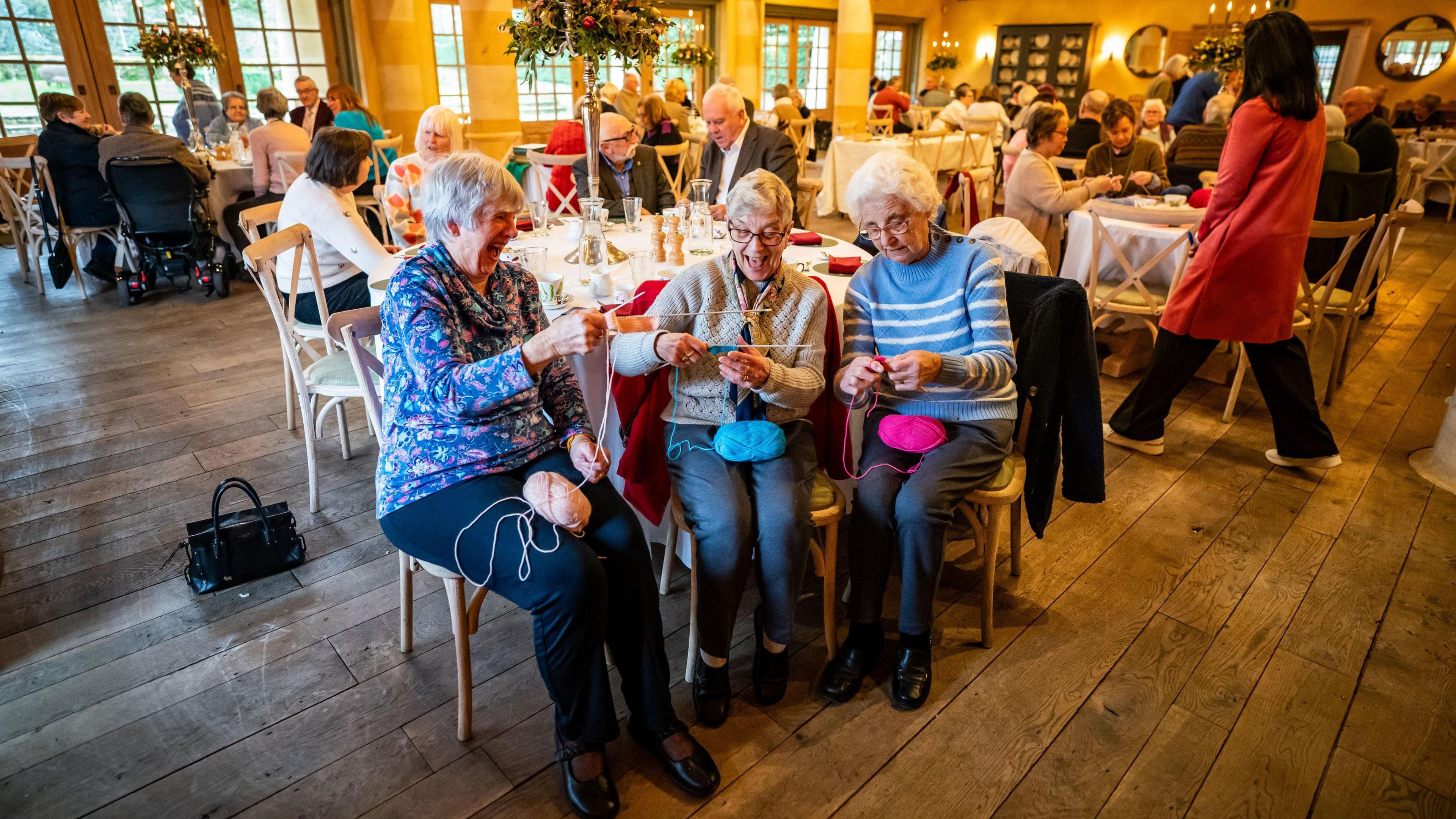 Three ladies knitting in a big hall at Highgrove with wooden floors and tall candlesticks.