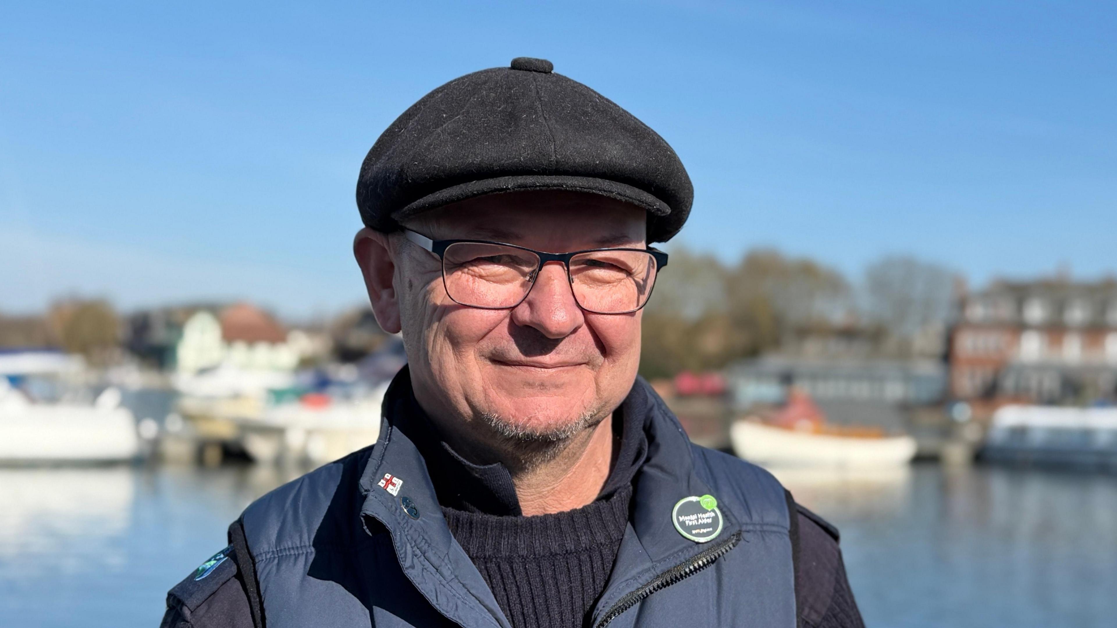 Paul Rice smiles at the camera. He wears a black cap with a blue gilet and navy jumper underneath. 