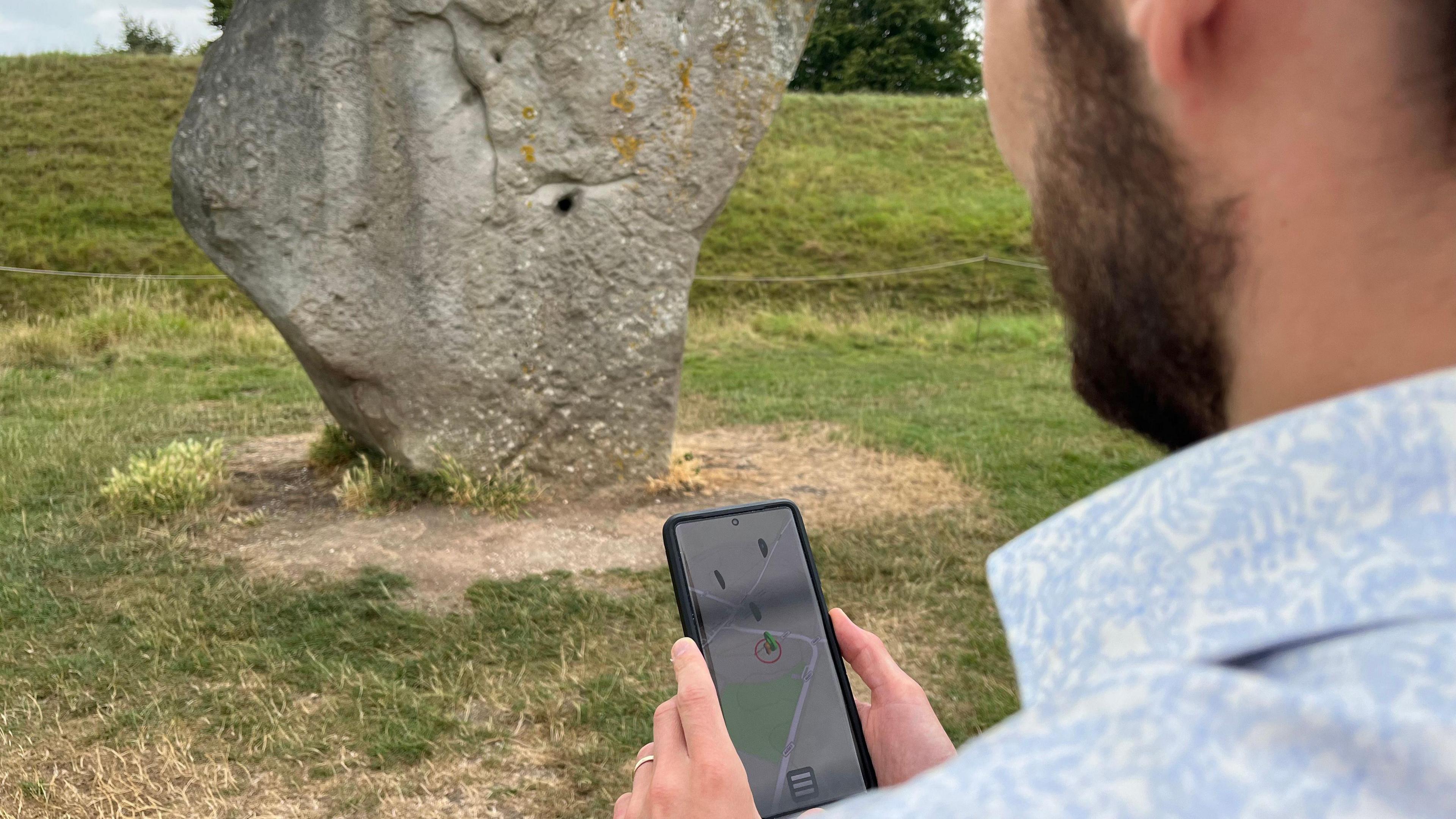Looking over the shoulder of a man with a beard, who is holding a smartphone. The screen shows a map with standing stones and a locator where he is standing. He is in front of one of the large standing stones at Avebury.
