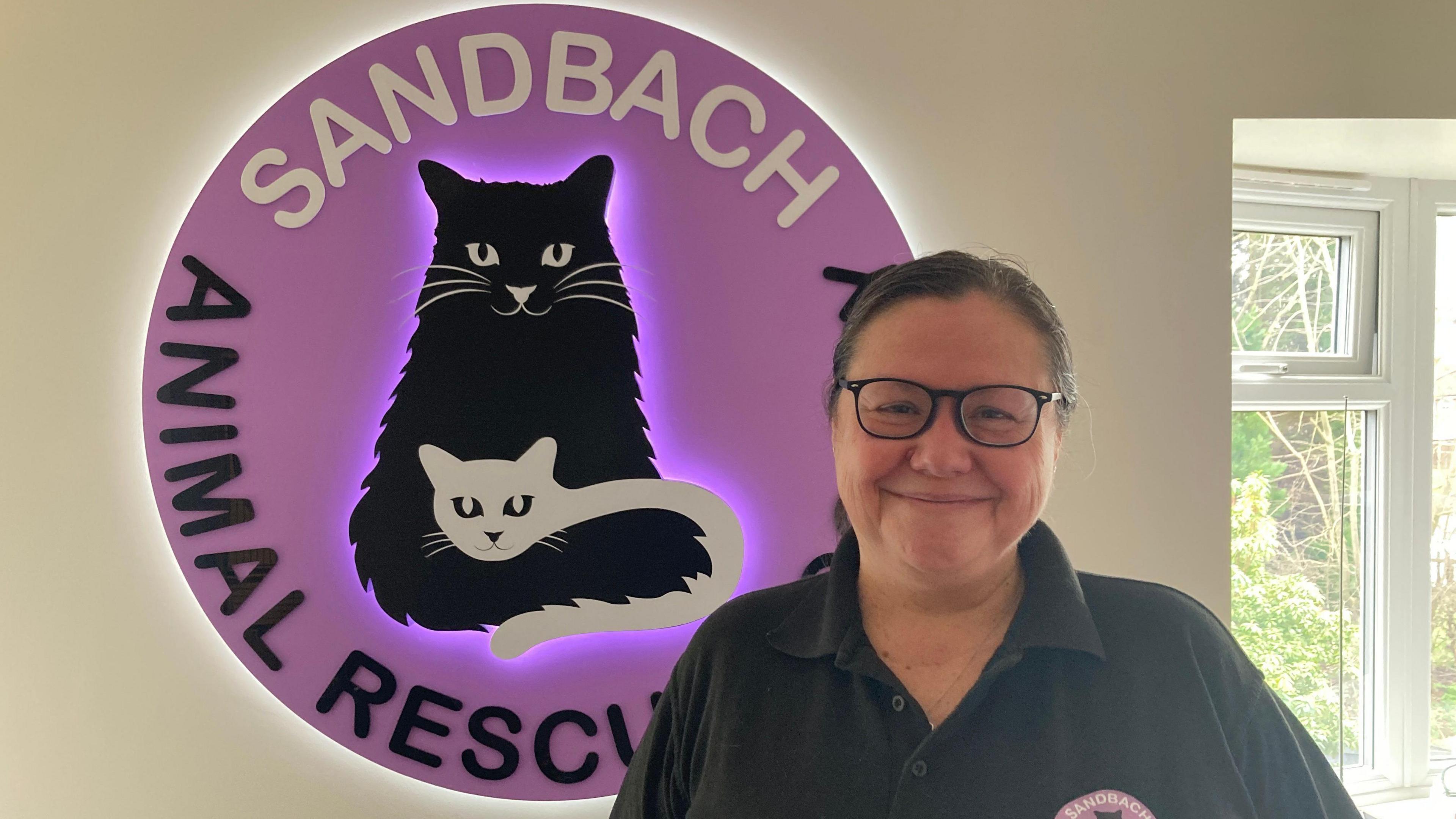 Samantha Moss, chair of trustees at Sandbach Animal Rescue, standing in front of the charity's logo. She is wearing a black t-shit and glasses. 