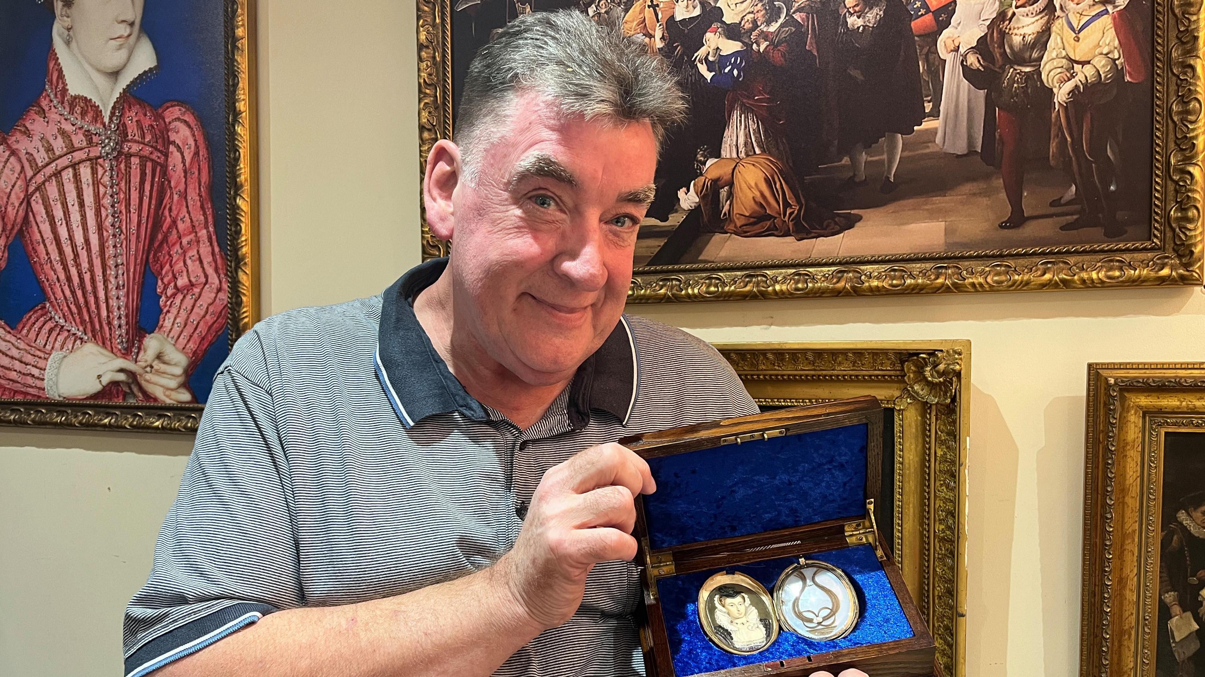 A man holding a lock of hair which is in a locket in a wooden box. There is a picture of a woman on the other side of the locket which is meant to be Mary Queen of Scots. He is wearing a short sleeved shirt and has short grey hair.