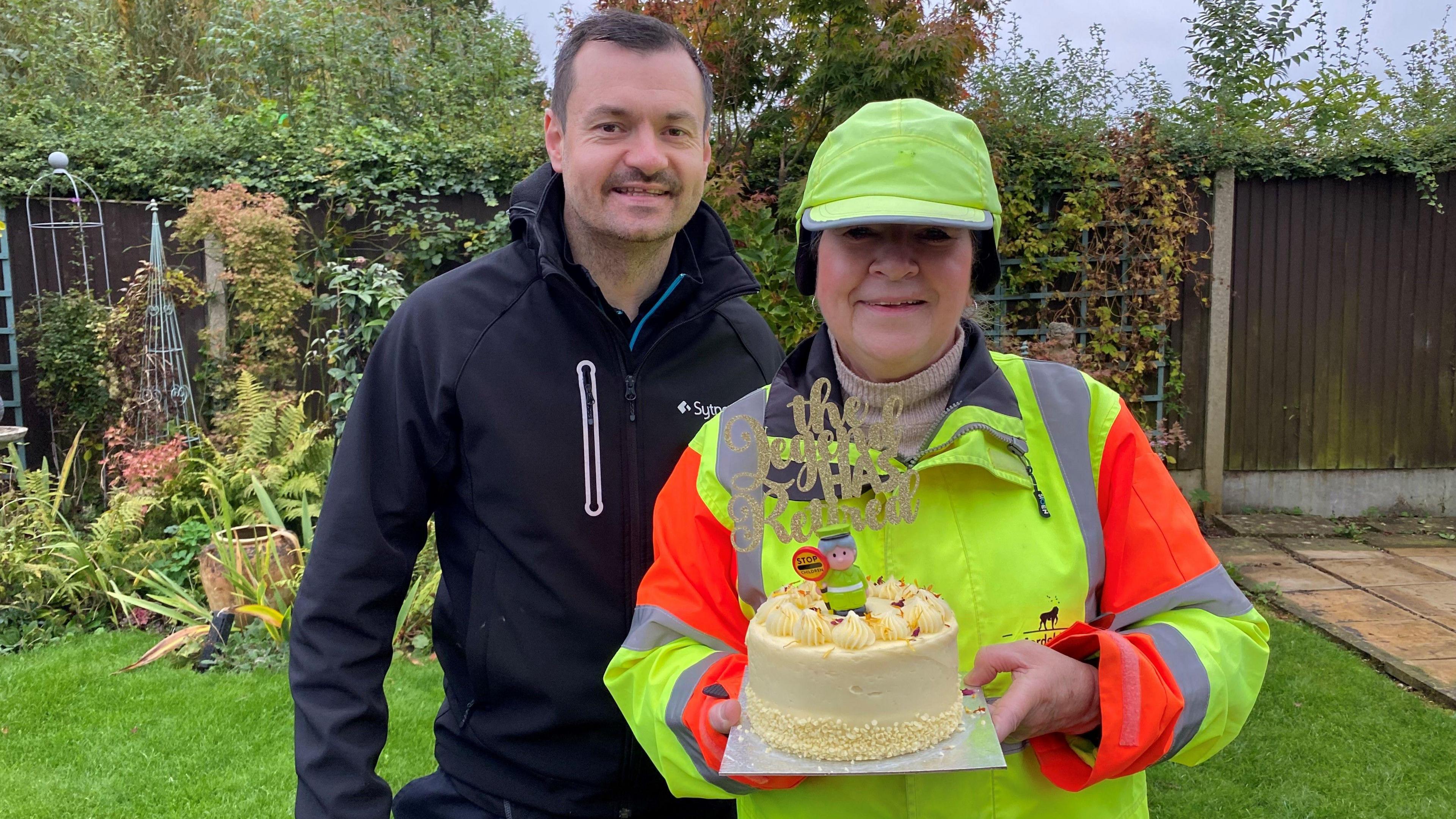 Julie Bennett, dressed as a lollipop lady, with a high vis hat and jacket, black ear muffs, standing beside her son, Rob Bennett. Julie is holding a cake that has a cake topper that says The Legend Has Retired, and a model of a lollipop lady. They are both smiling and looking straight at the camera. Rob is wearing a dark jacket and has a slight beard.