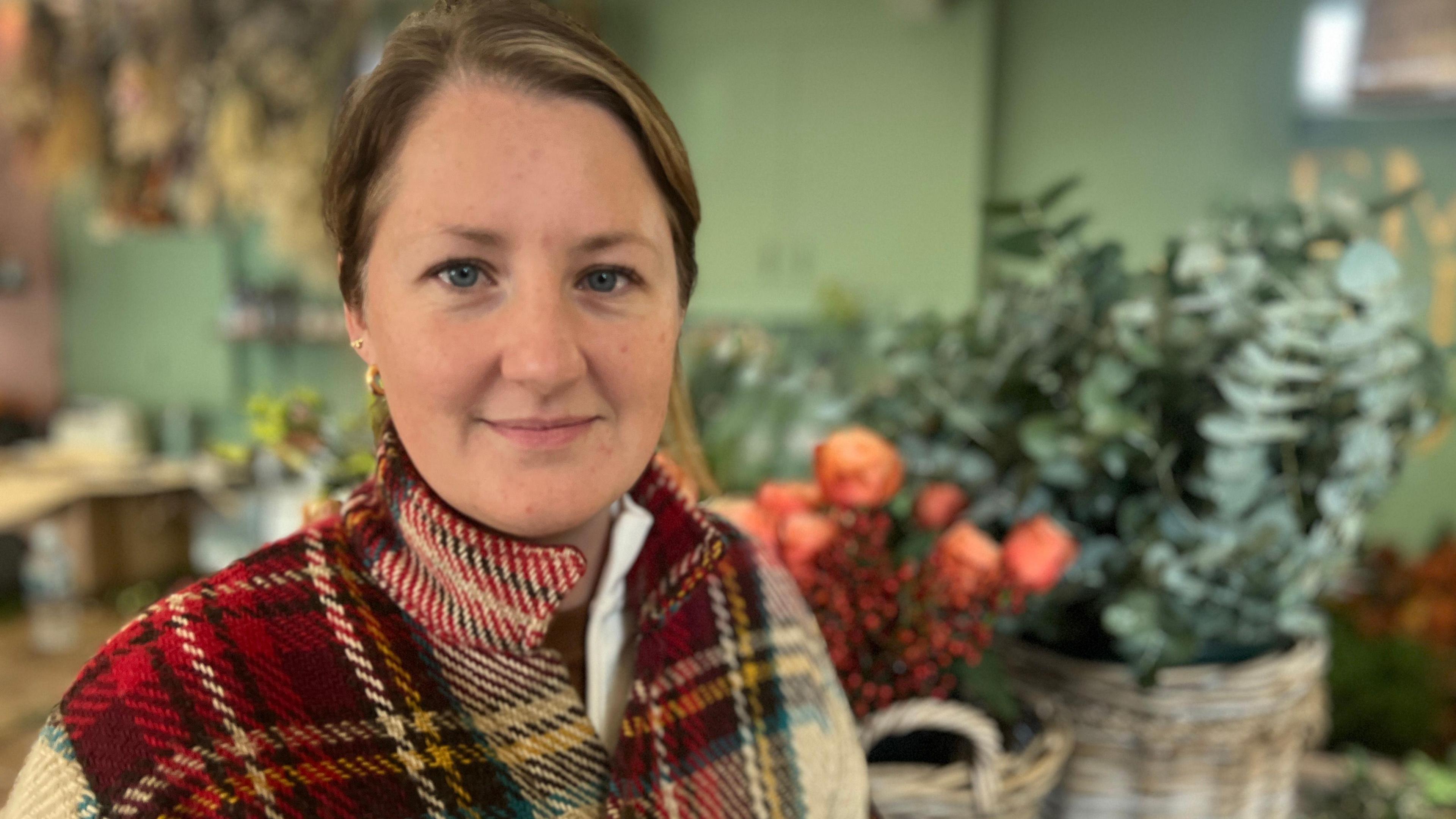 Emily Watson looking into camera in front of flowers in her shop. She has tied back blonde hair and is wearing a tartan-style jacket.