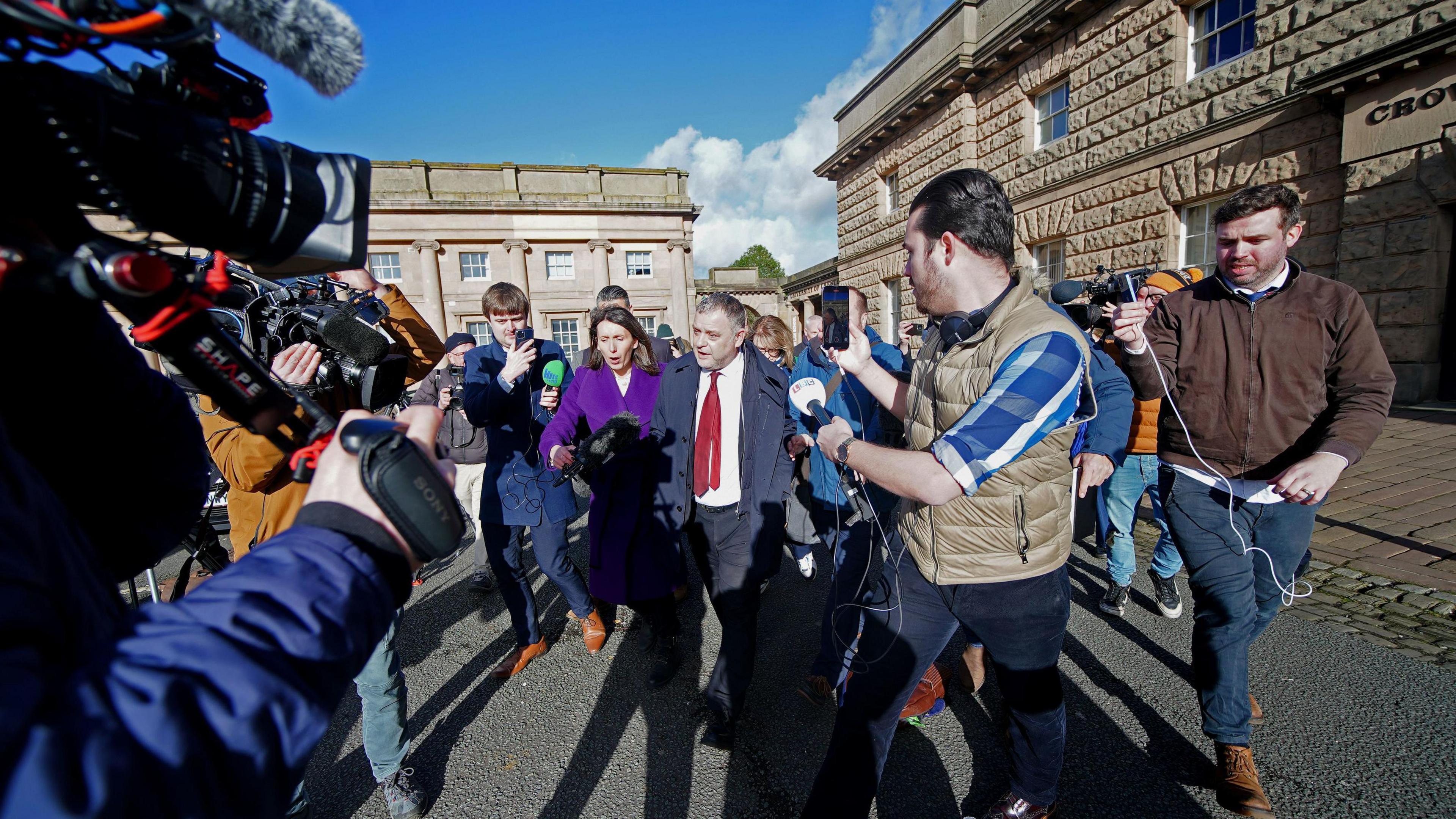 Media surround MP Mike Amesbury as he leaves court. Reporters are holding microphones towards the MP, who is wearing a dark blue jacket over a white shirt and red tie.