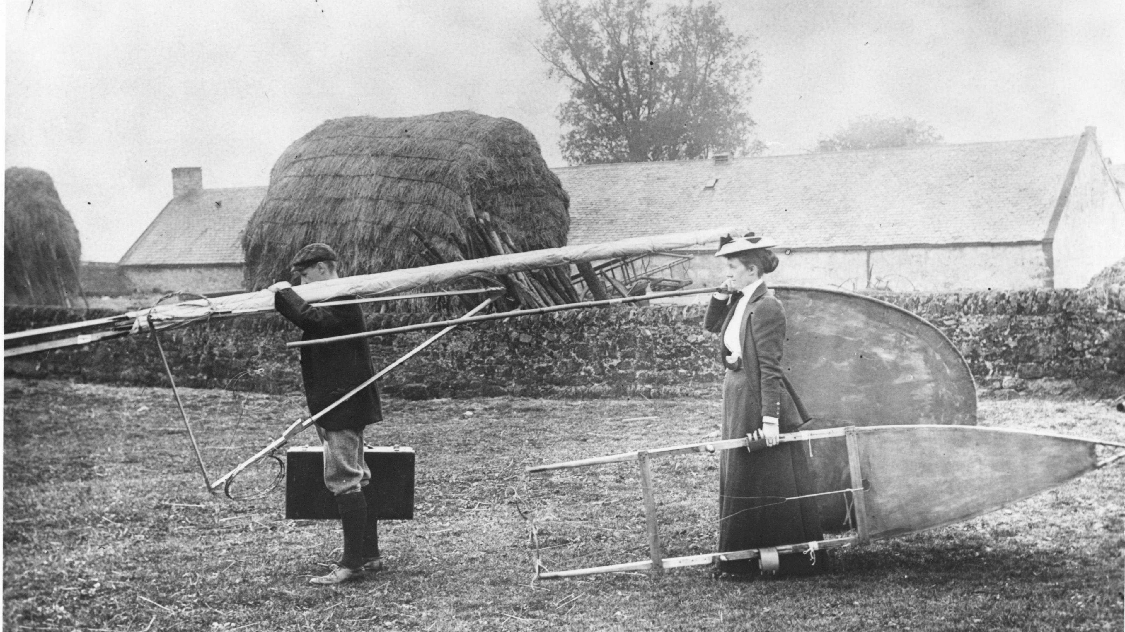 A black and white image of Percy and Ella Pilcher, both in Victorian-style dress, carrying the dismantled Bat glider through a grass area. A house or barn building can be seen in the background behind a small stone wall.