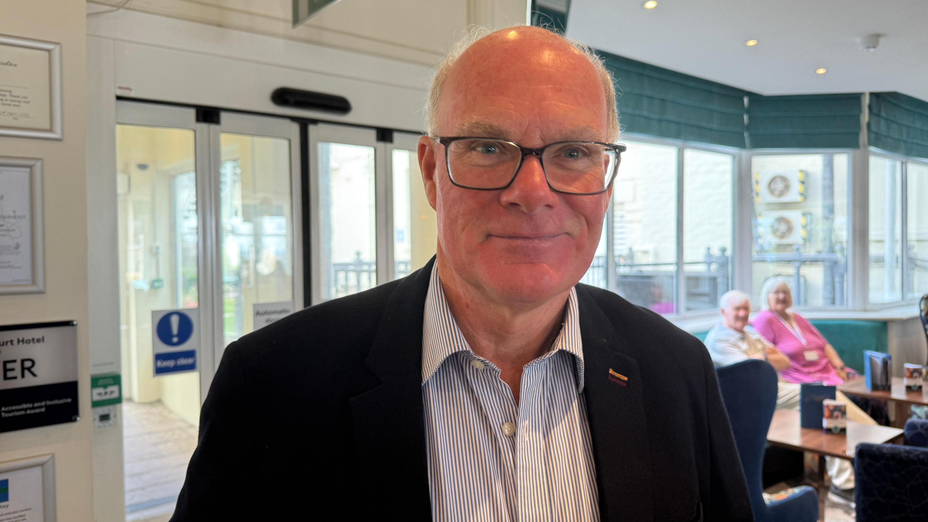 A man stood wearing glasses with a striped shirt and black blazer.