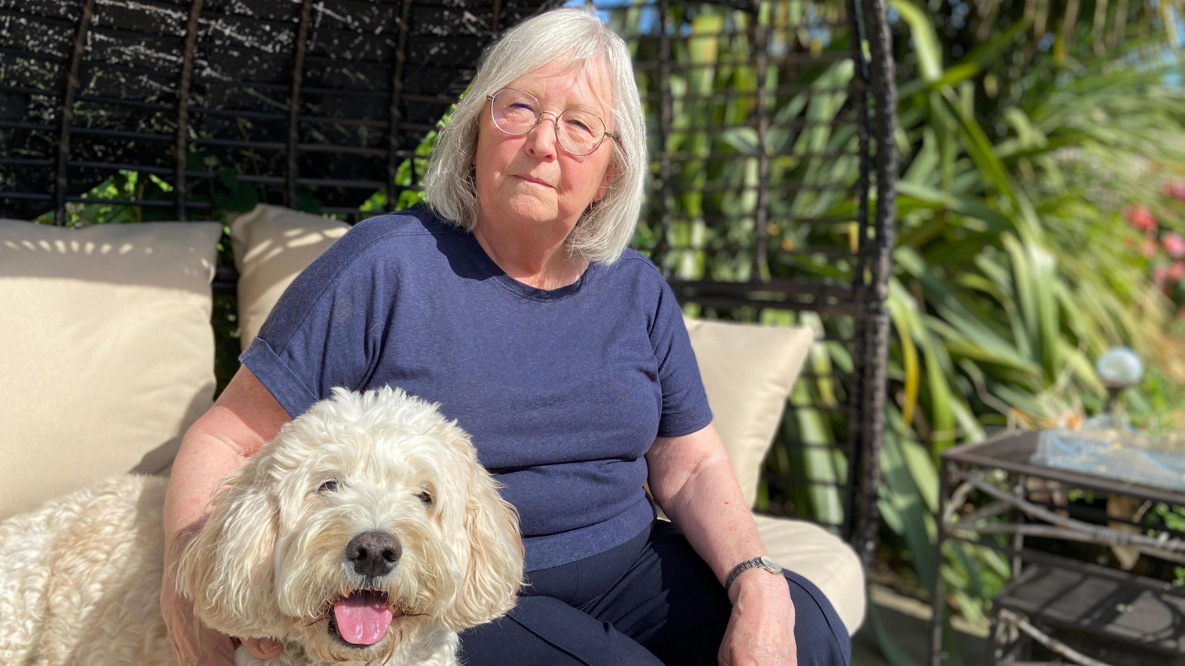 Avril is sitting outside in a blue top. She is sitting on a black wicker chair. Beside her is Dougie who is a white dog. 