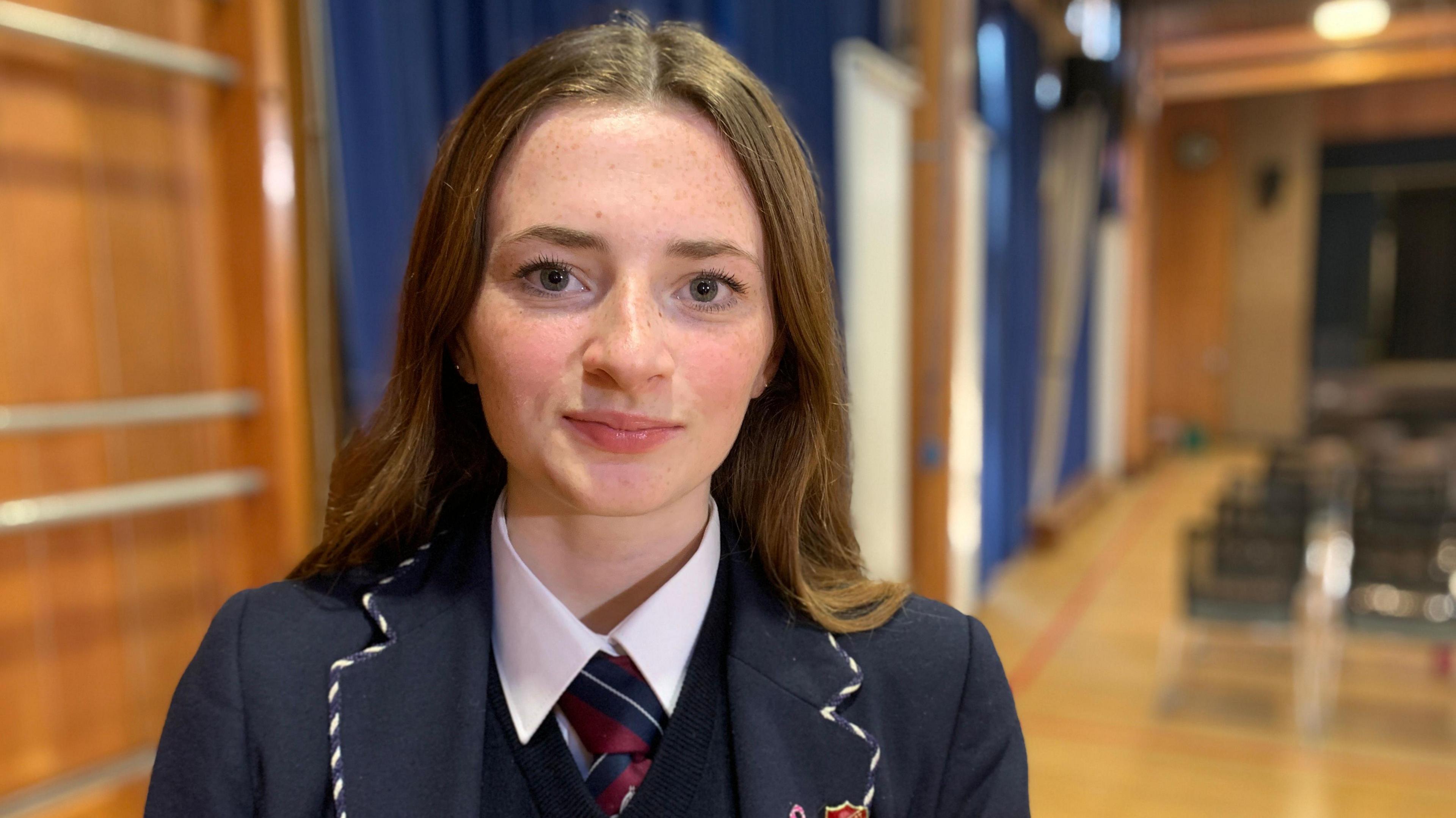 Francesca,  who is wearing a navy school uniform, has light long brown hair, and dark brown eyes, looks at the camera with a neutral expression