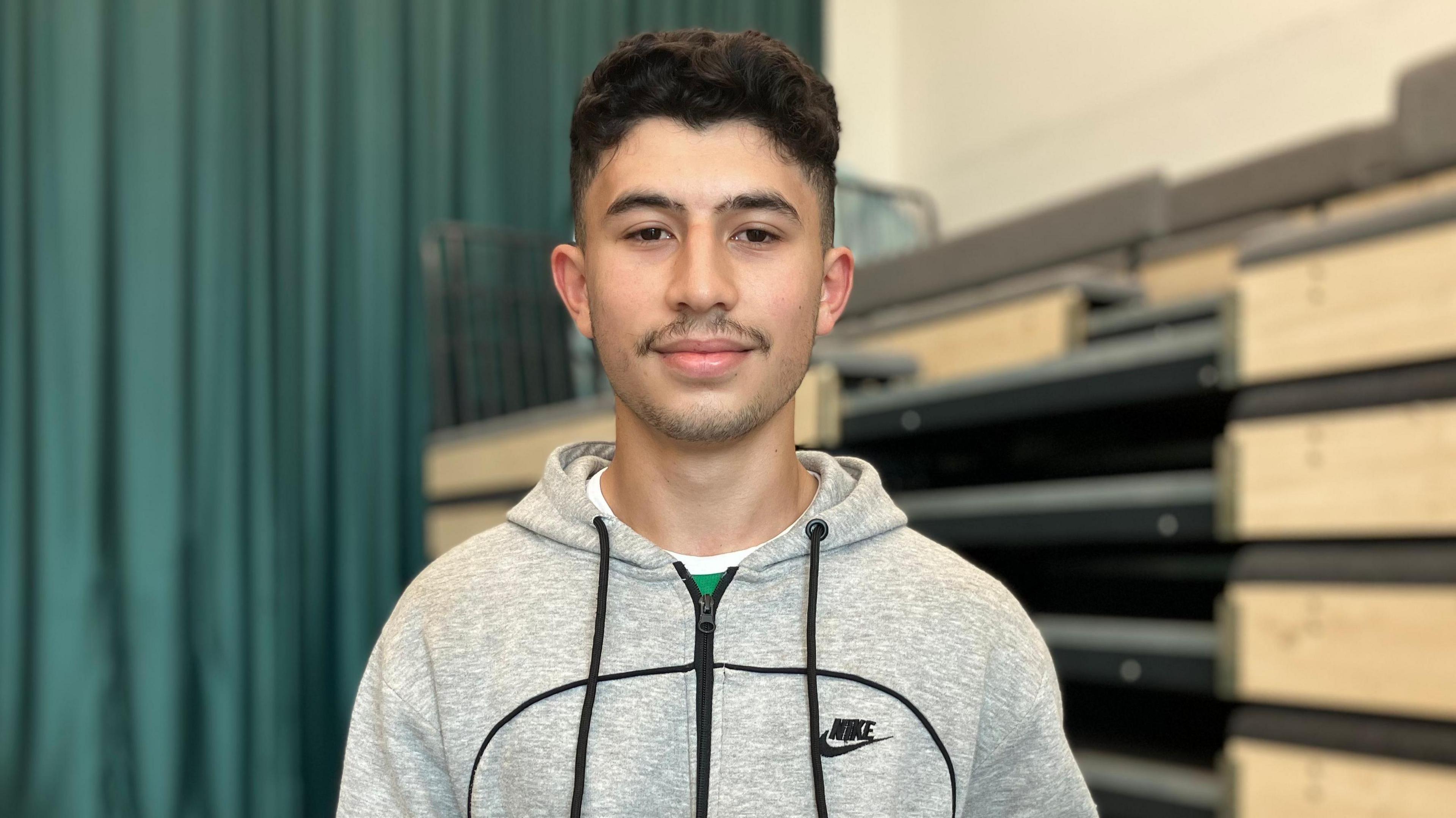 Mohamed Farhoud standing in the school hall wearing a grey zip up Nike hoodie and smiling at the camera