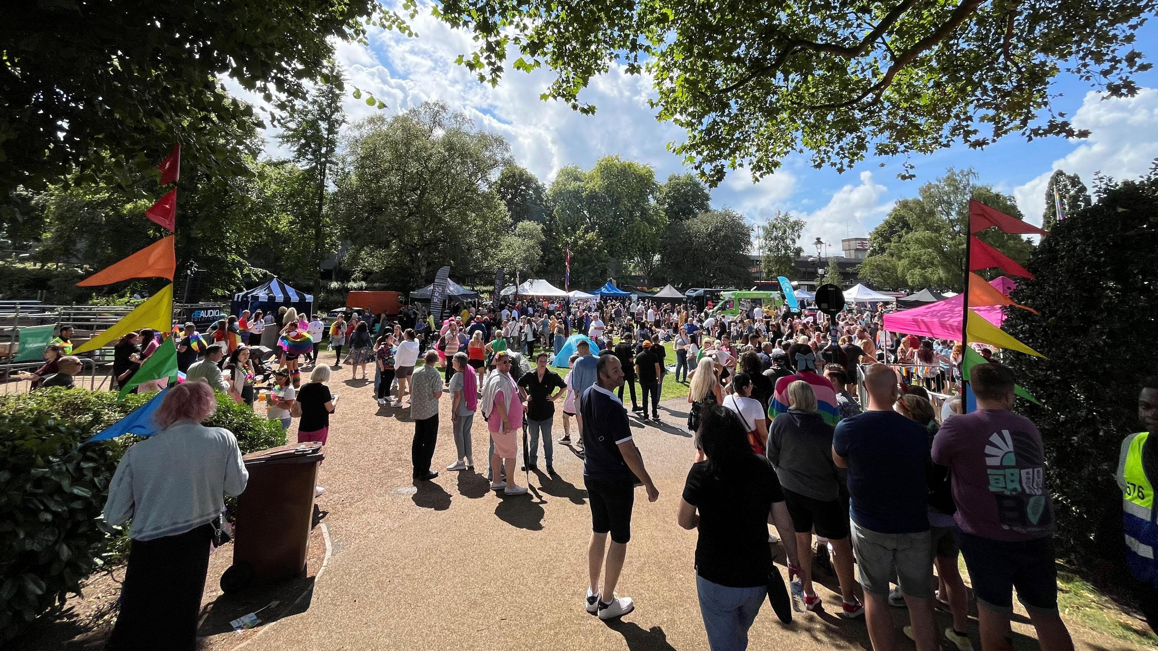 A large crowd of people standing in a park with gazebos and stalls visible in the background. Rainbow flags are on posts either side of the entrance.