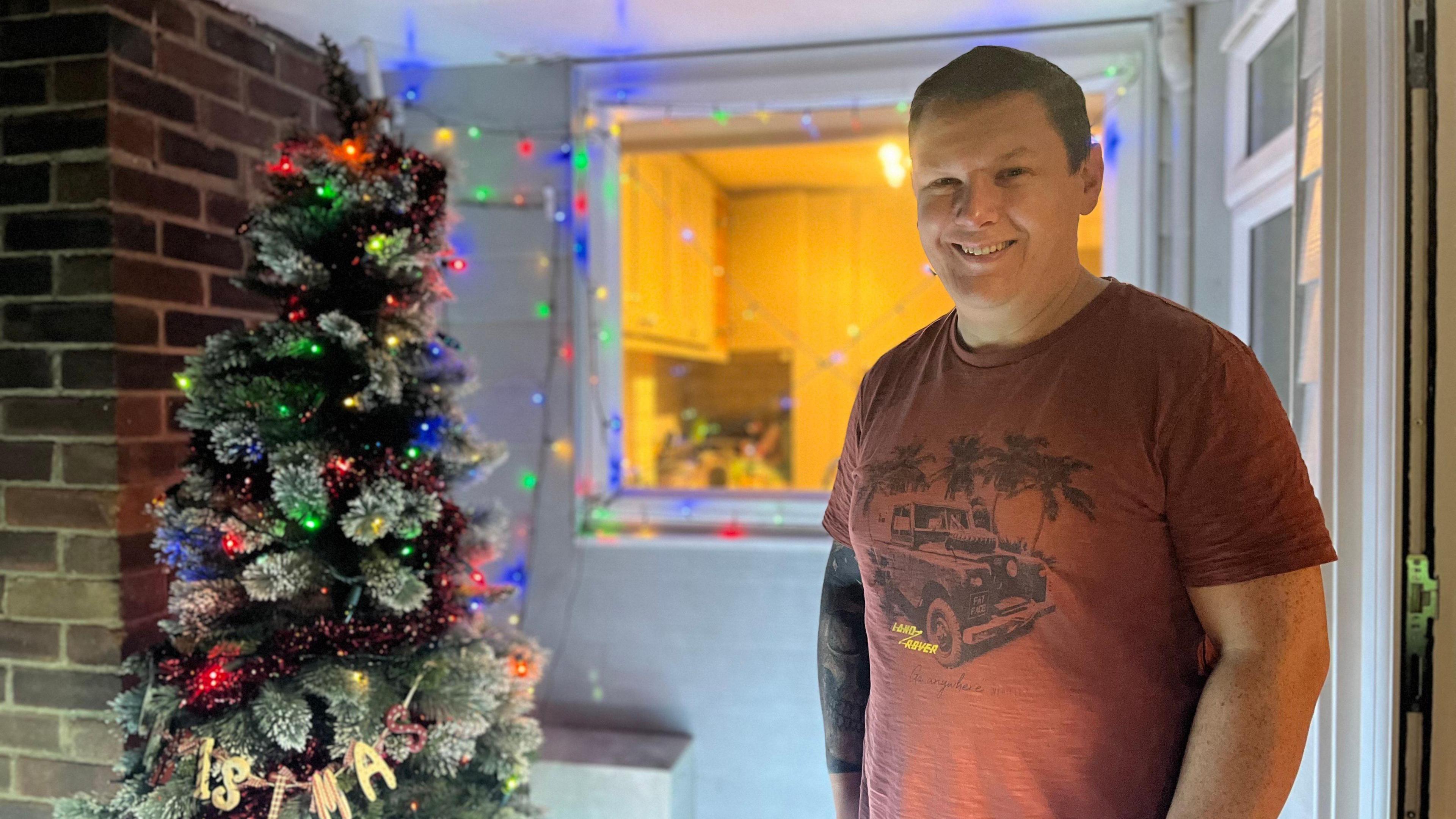 Sam Aylward stands in the doorway of a house with a Christmas tree to his side. There are lights on the tree and around his window.