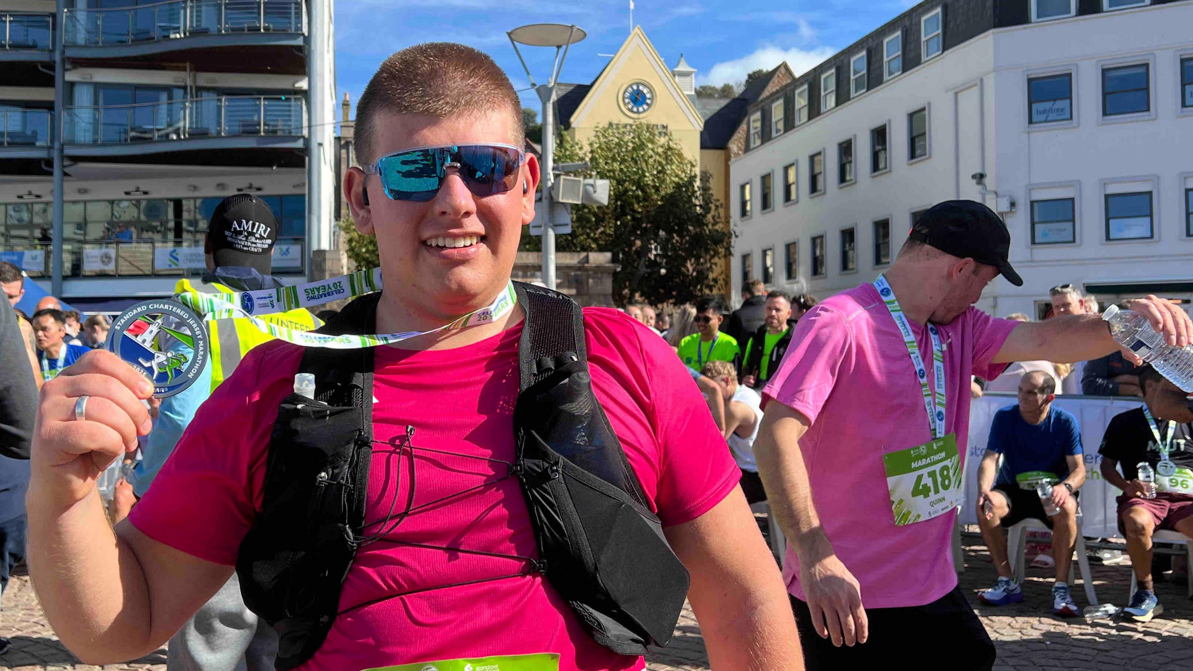 Ryan is wearing sunglasses and a bright pink shirt with black straps, holding up a medal. The green race bib reads 559. Other participants and buildings, including one labelled 'The Royal Yacht', are visible behind.