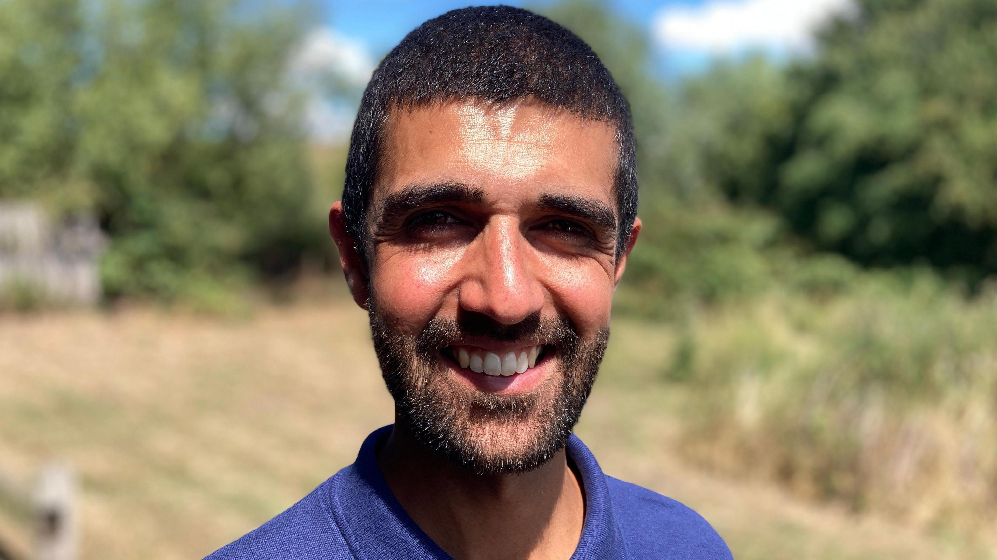 Ajay Tegala, who has short black hair and a short black beard and is wearing a blue top, smiles at the camera. He is outside and there are green shrubs and trees in the background, which is out of focus.