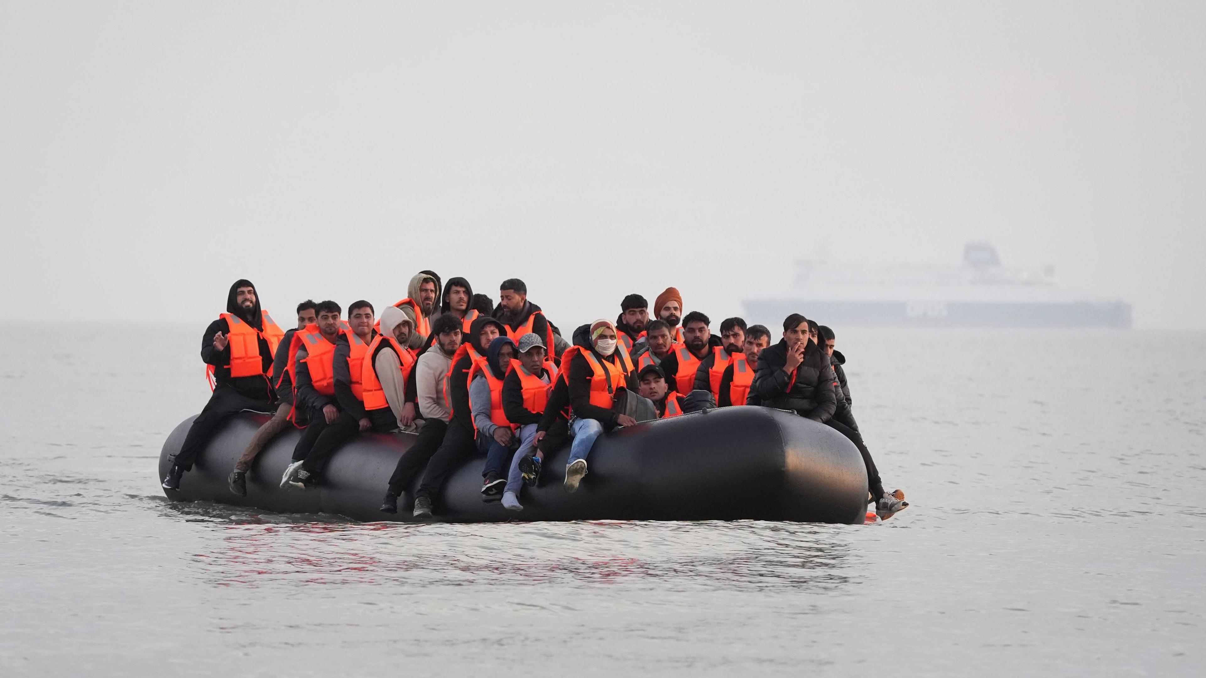Migrants sit in an inflatable boat as they travel across the English Channel. They are wearing orange life jackets.