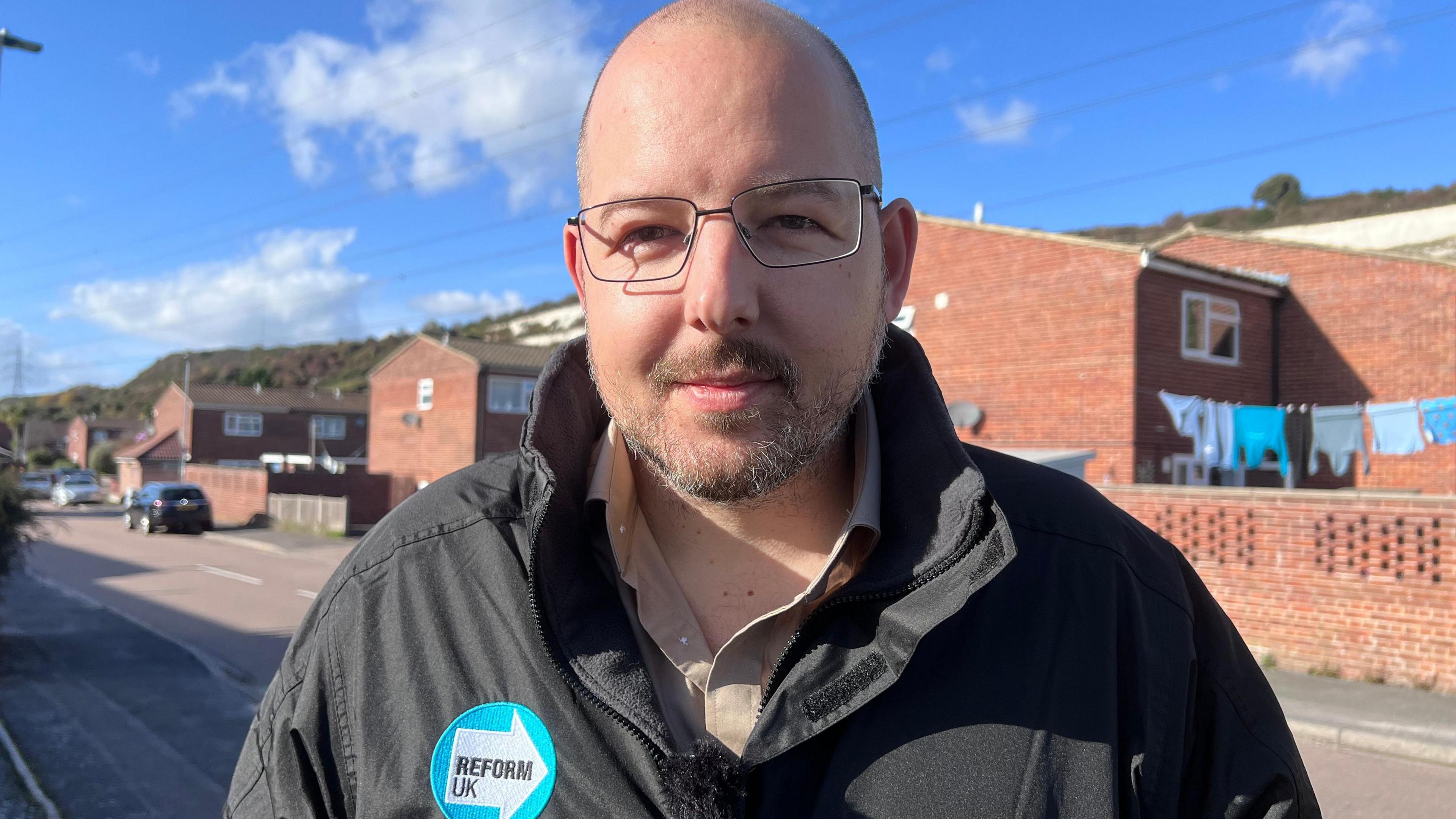 Bald man with glasses wearing a Reform UK jacket stands in front of houses in Portsmouth