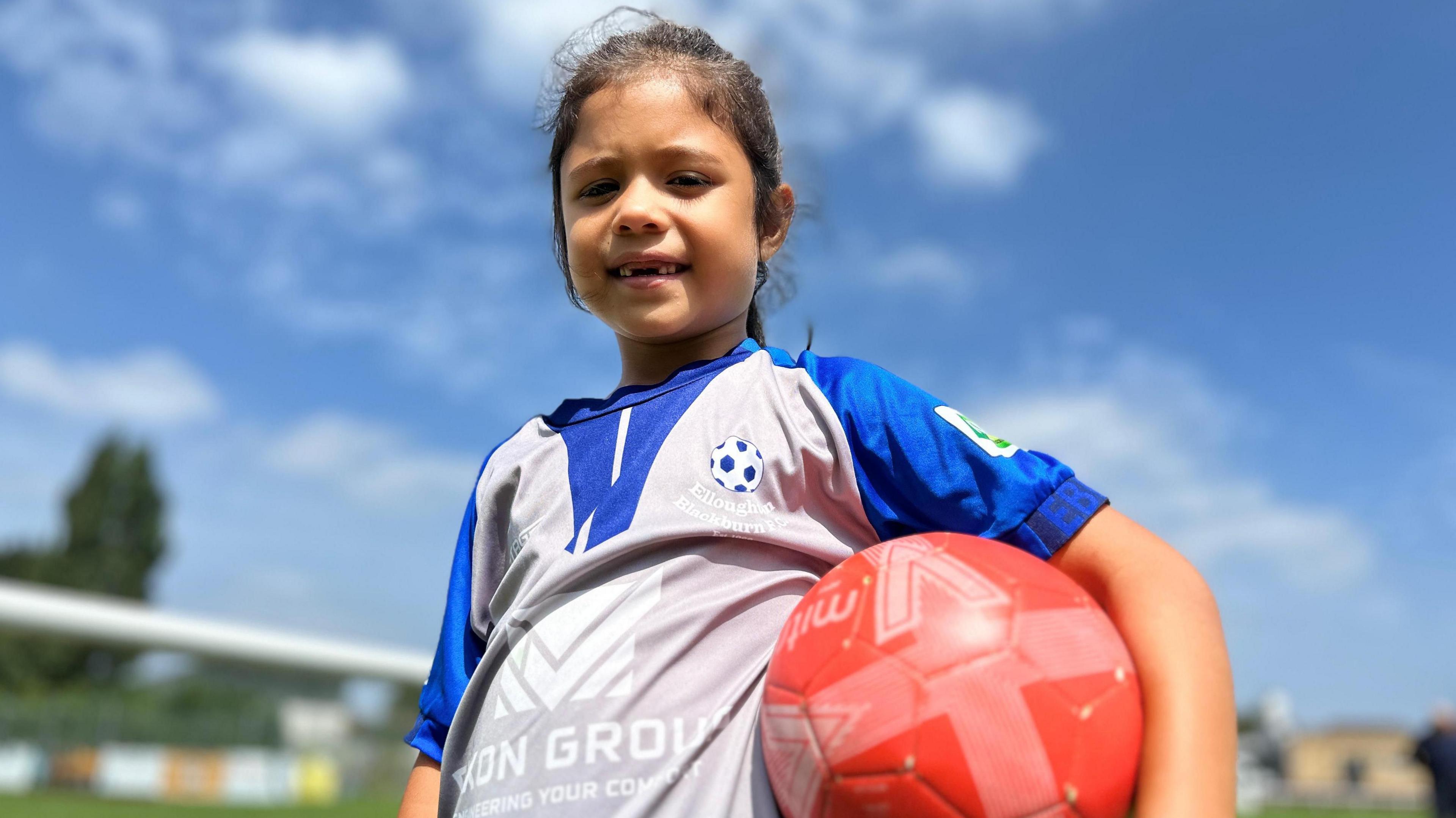 Young girl with dark hair and wearing a blue and grey football shirt. She is holding a red football.