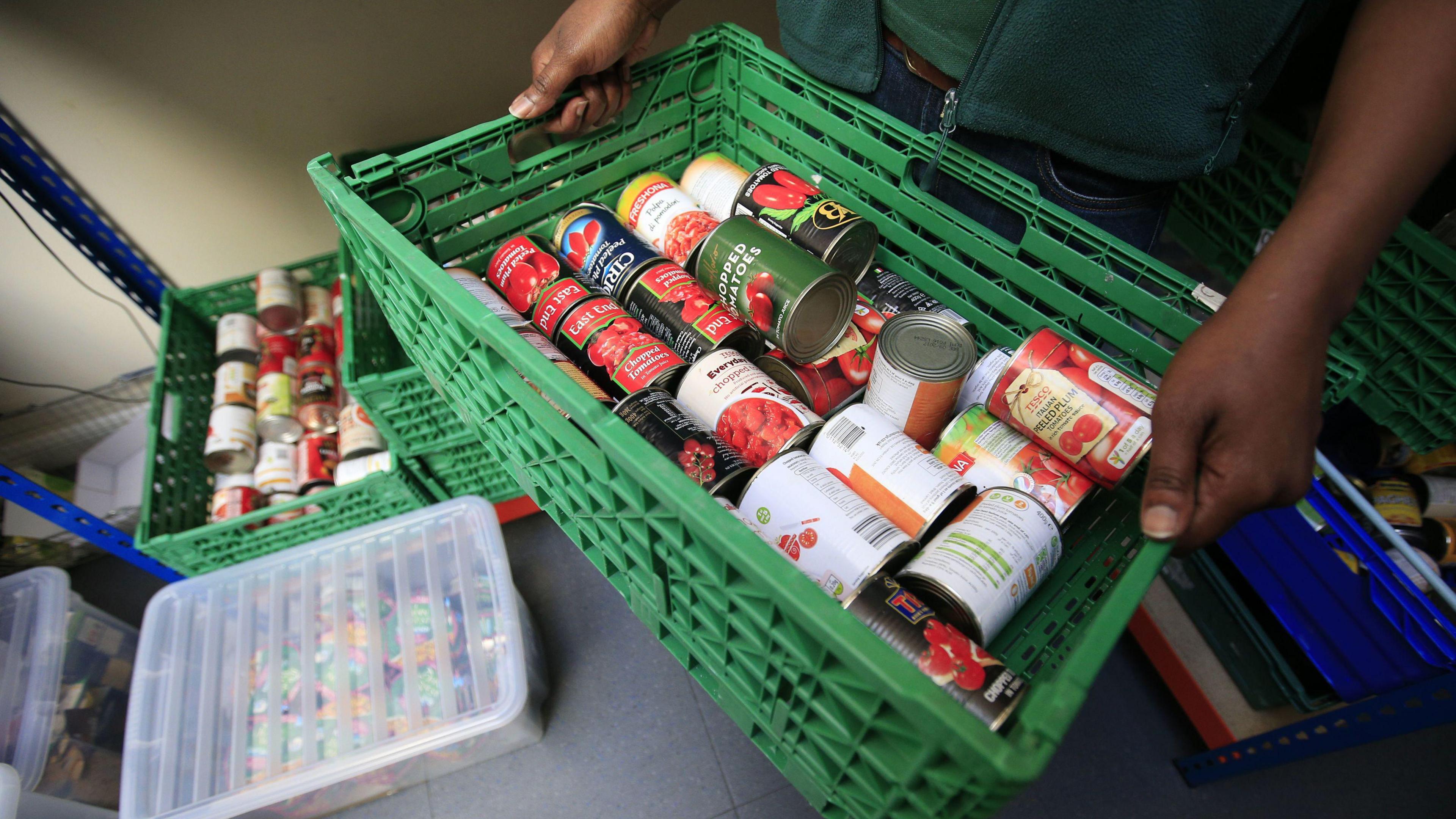 A green crate containing tinned foodstuffs is held by a person wearing a green gilet. There are more crates of tinned food on the floor.