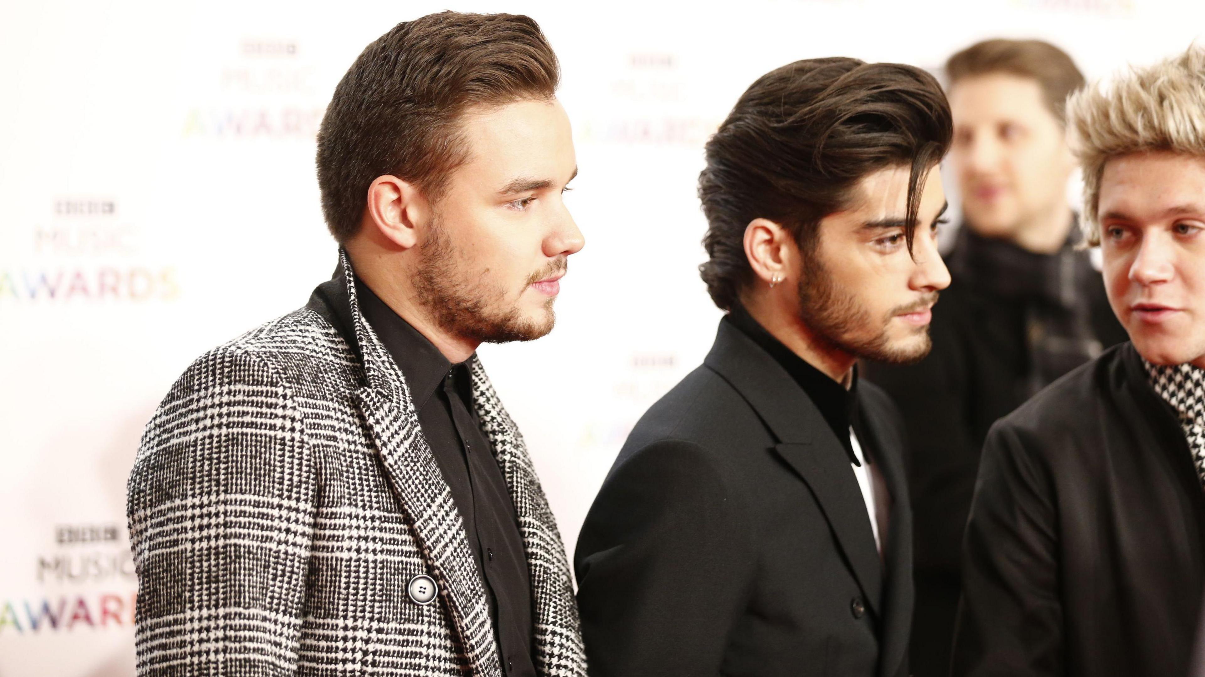 From left to right: Liam Payne, Zayn Malik and Niall Horan at the BBC Music Awards in 2014. They are all dress in dark suit jackets and shirts and have their hair slicked back. Payne and Malik are looking to the right. Horan is speaking to Zayn. 