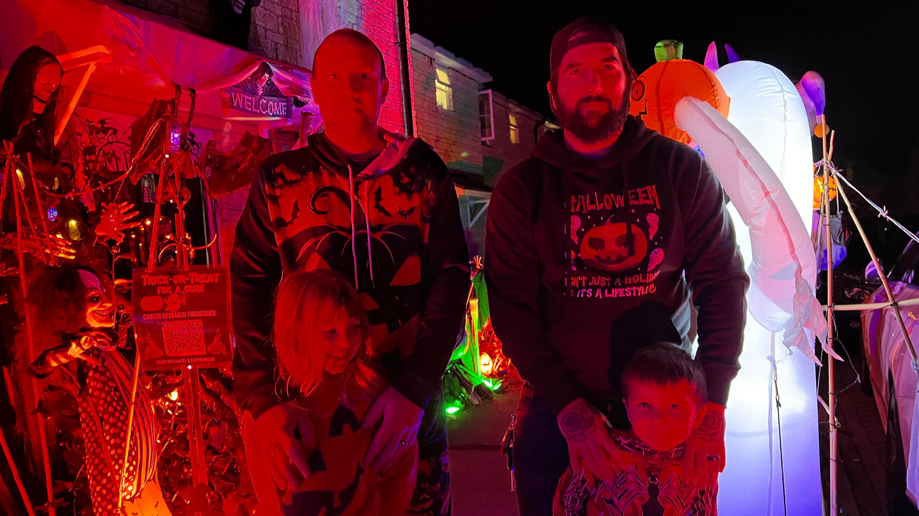 Two men, both in Halloween jumpers, with two young children in front of spooky inflatables and props.