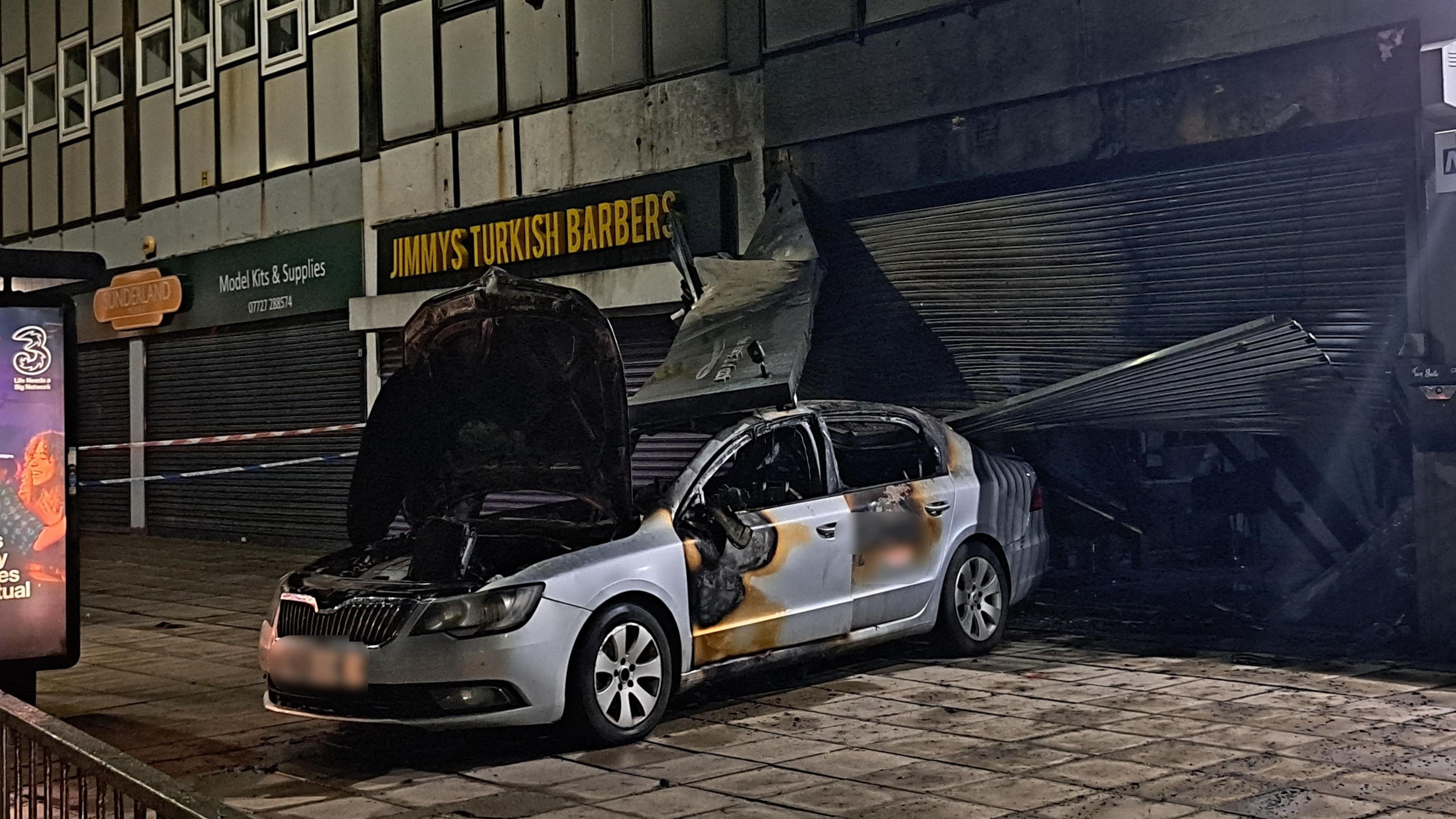 A burnt out silver car on a pavement in front of a shop. The shutters of the shop are mangled