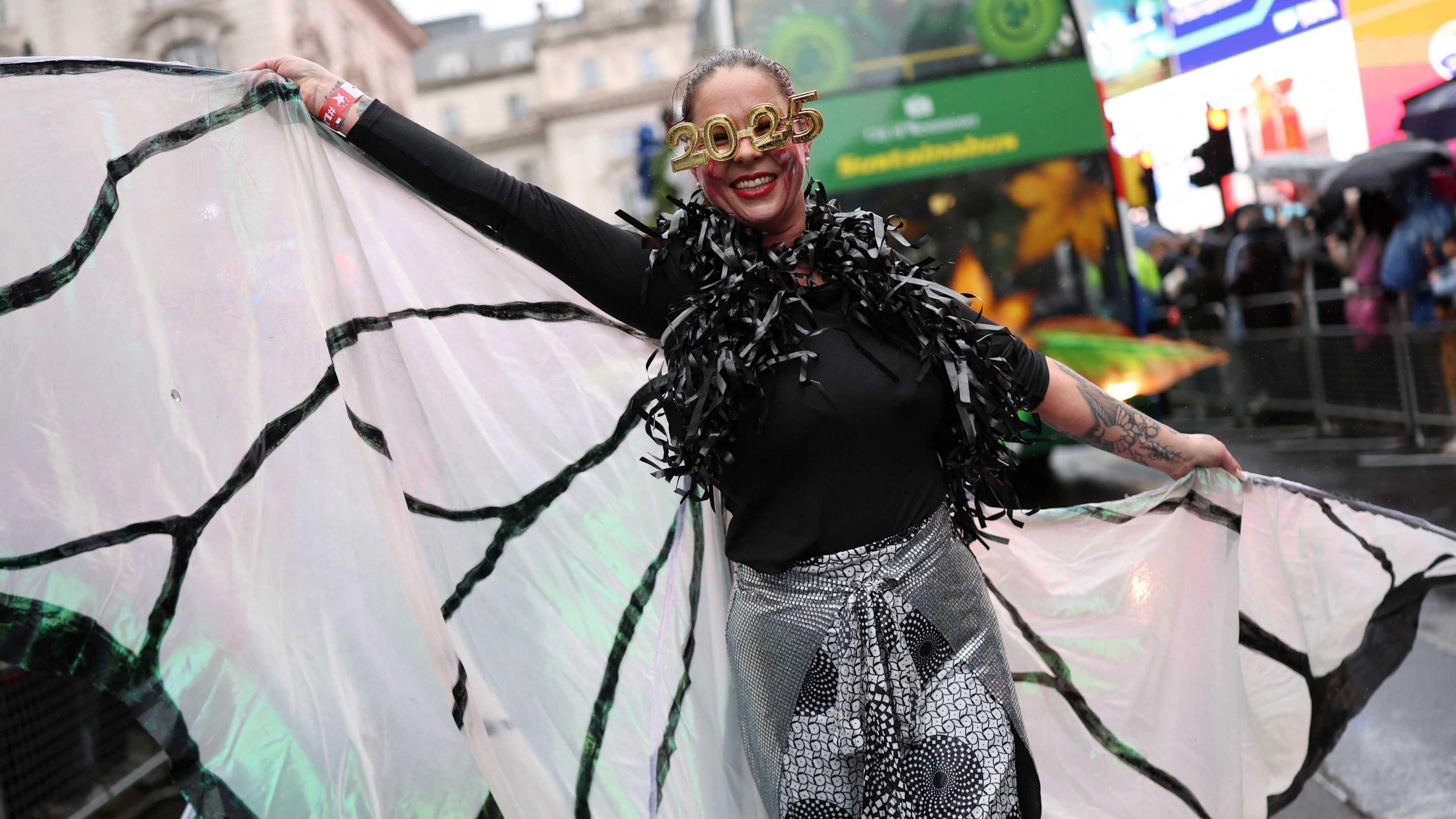 A woman participant in the street in Piccadilly Circus wearing a black feather boa and a black top, novelty 2025 glasses and a flowing white and black wings. 