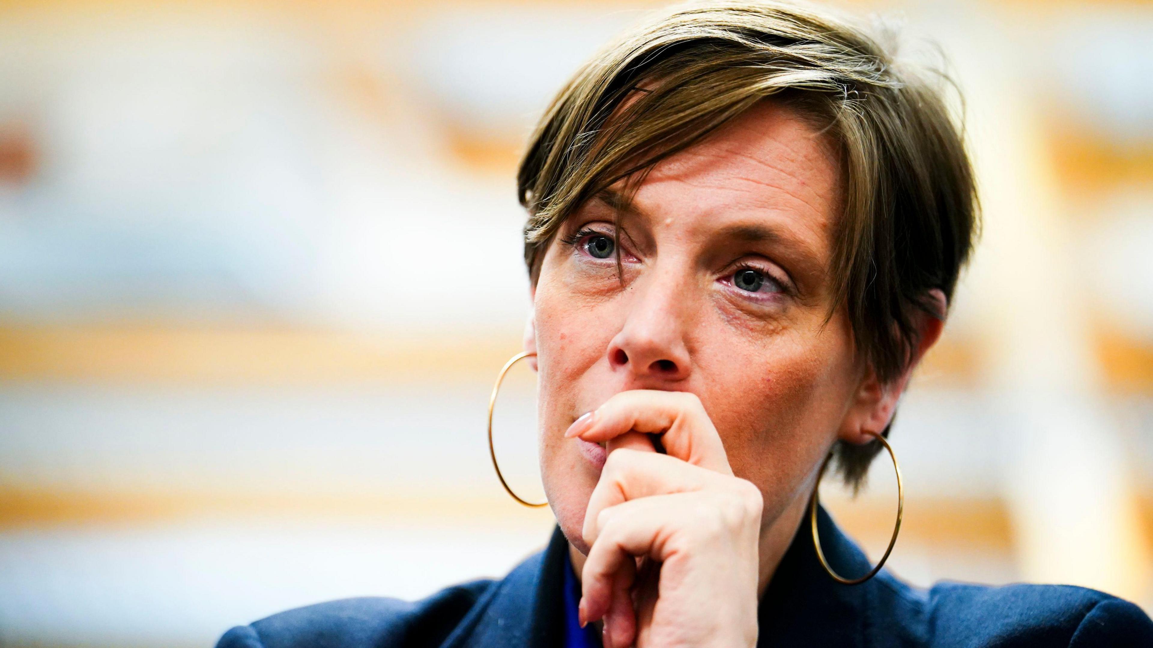 Government minister Jess Phillips sits holding her hand in front of her mouth looking thoughtful. She has shortish dark brown hair streaked with lighter brown and is wearing large gold hoop earrings.