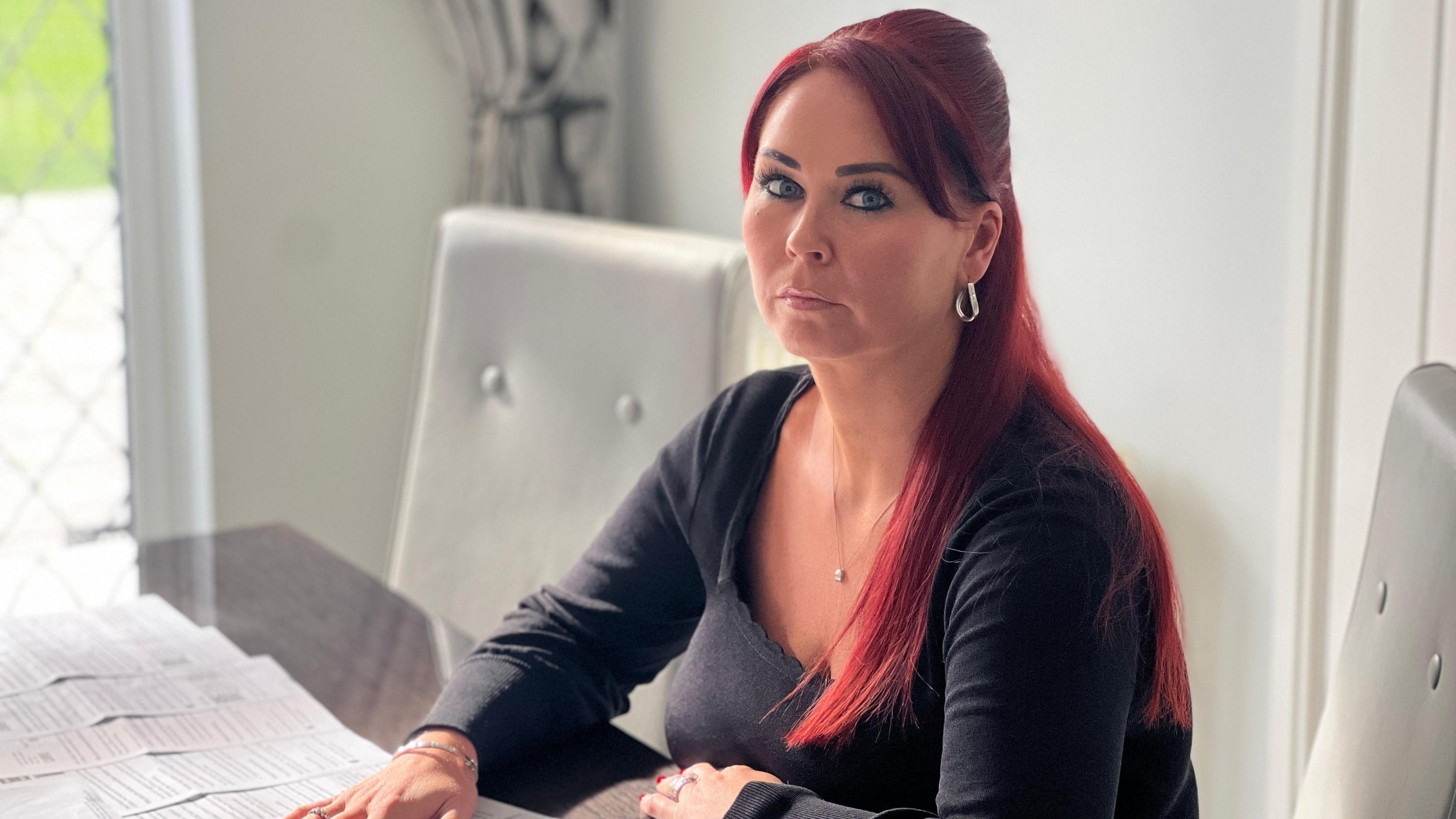 Rachel Canning, sits at her dining room table with her hand on a "removal notice". She has red hair, and wears a plain back top, silver hoop earrings and a small pendant necklace.