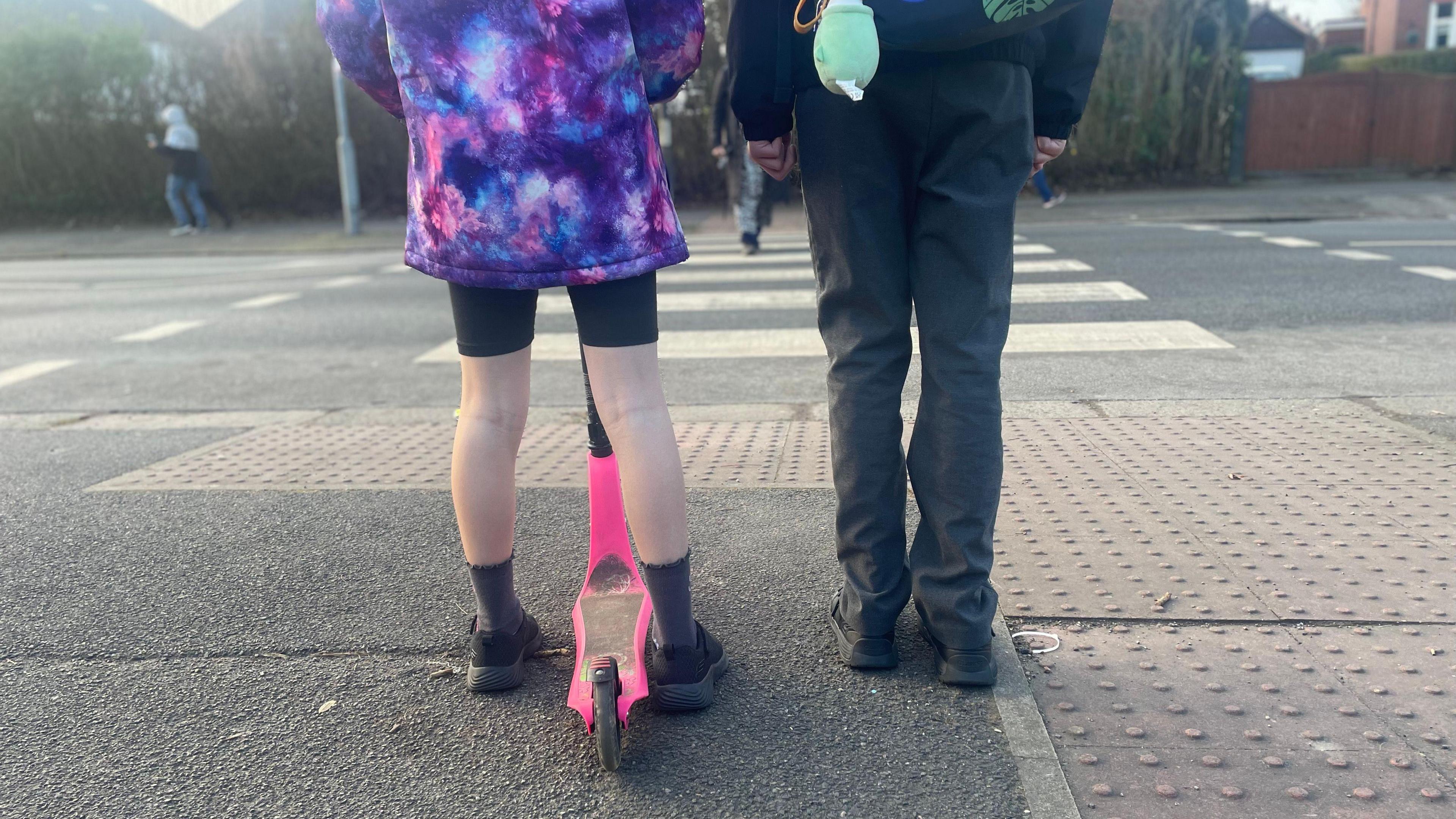 The back legs of two school children, one with a scooter between their legs, standing at the side of the road with a zebra crossing in front of them.