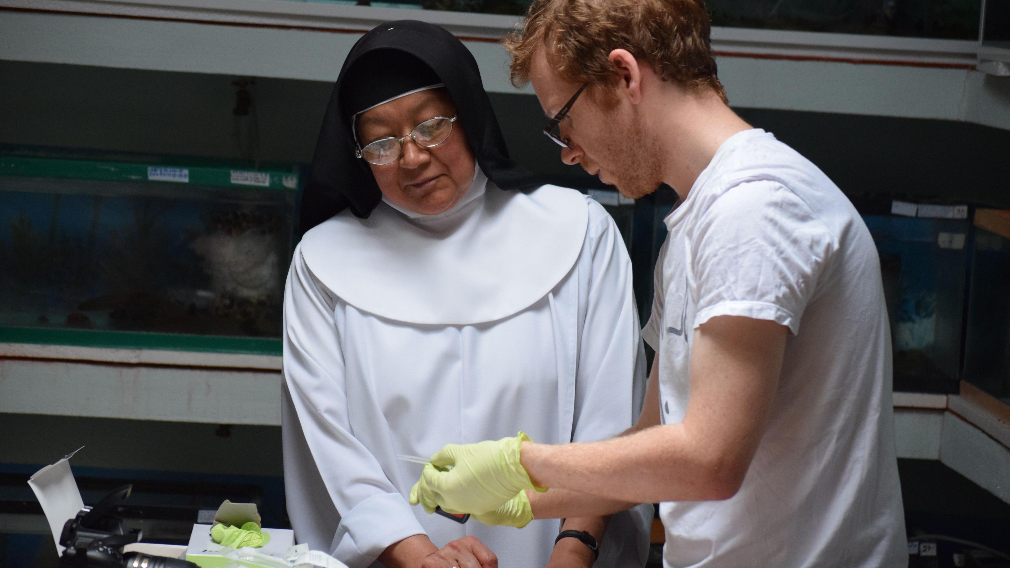 A nun and a scientist who are in a laboratory. The scientist has red hair and is wearing a white tshirt and green gloves. He is holding something in his hands that the nun is looking at.