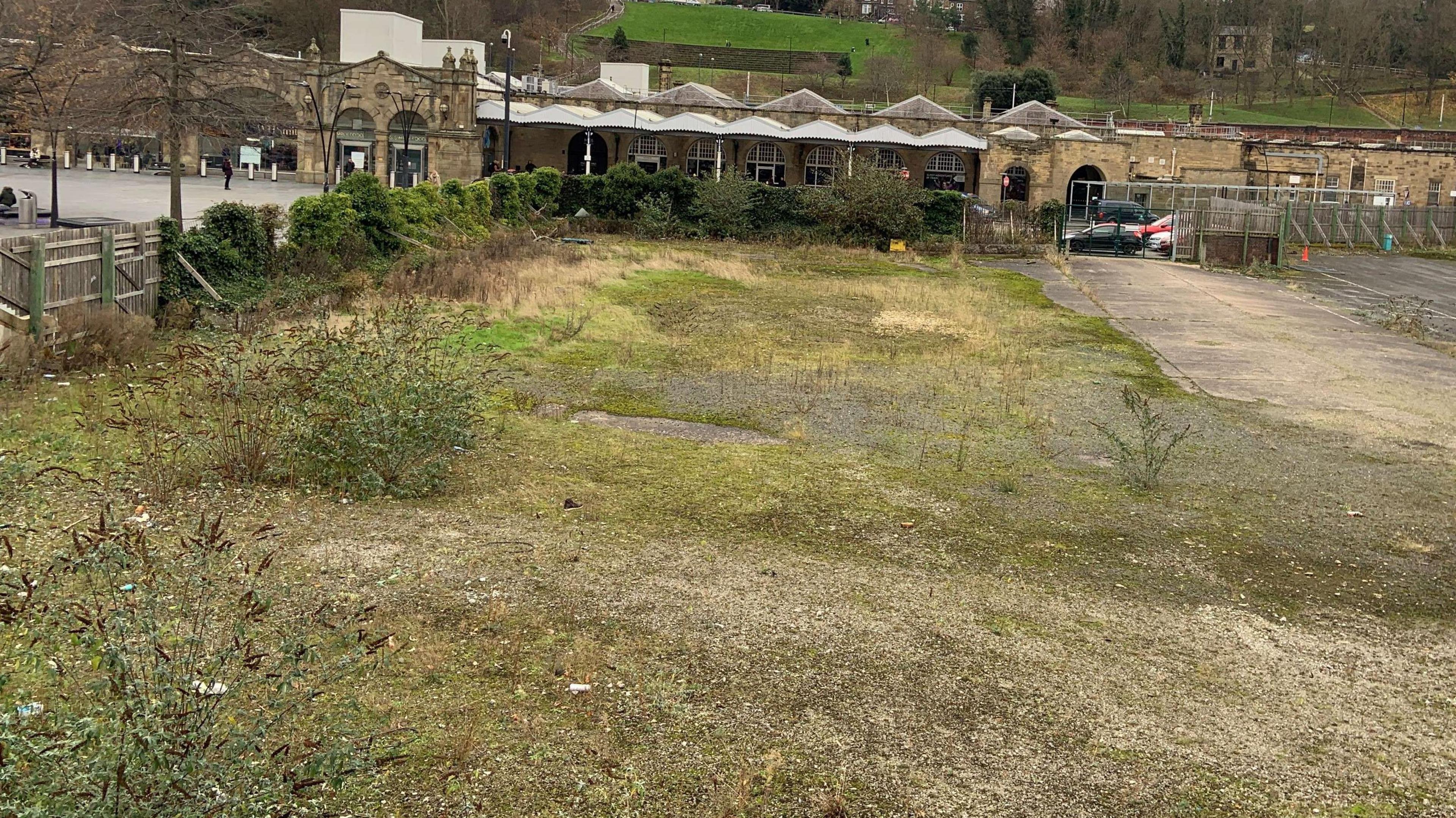 Photo shows derelict land by Sheffield railway station. There is some scrubland grass and a metal fence around part of the site