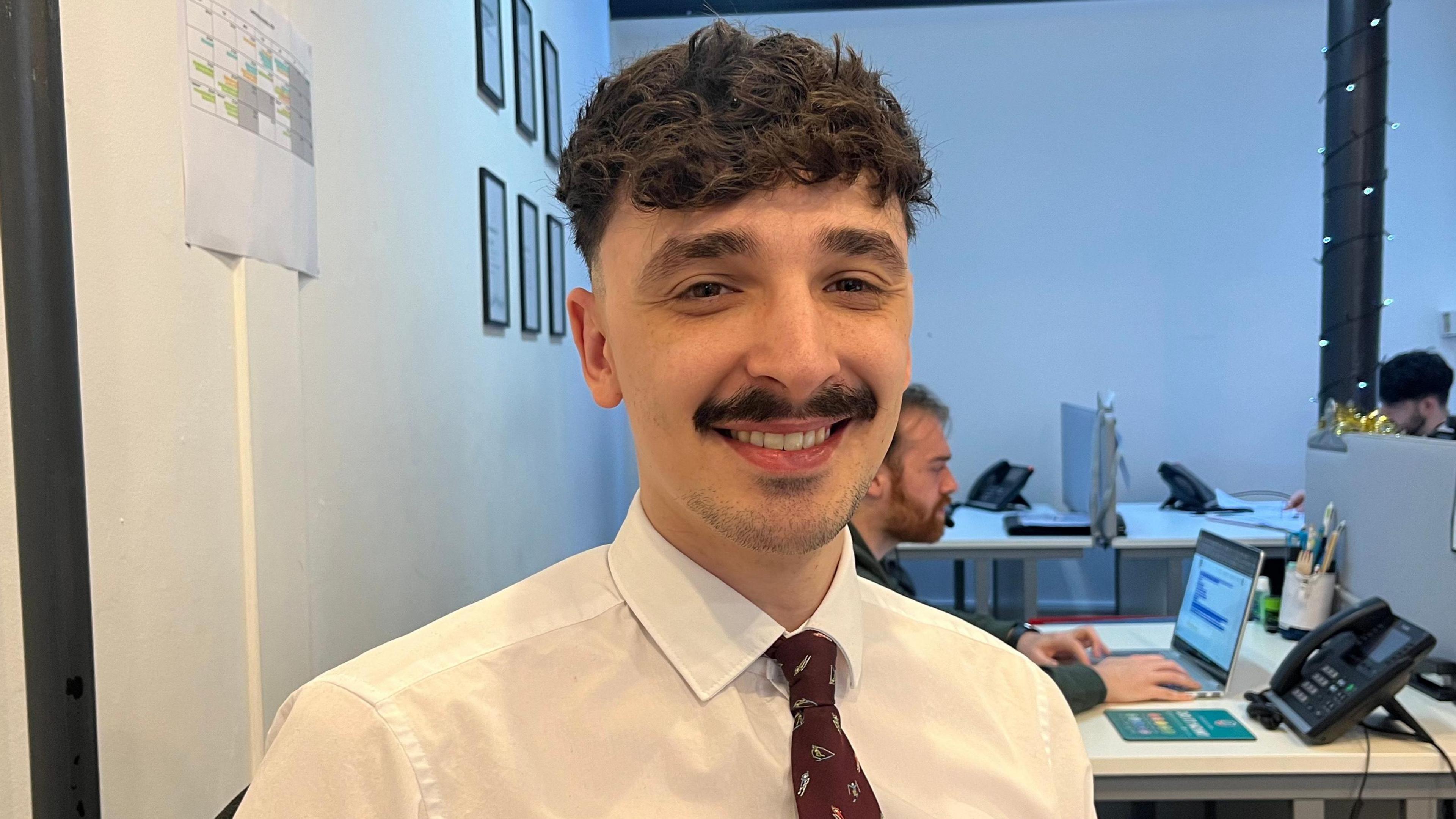 Head and shoulders of Kyle Butterworth who is wearing a white collared shirt and red tie. Computers and desks can be seen behind him.