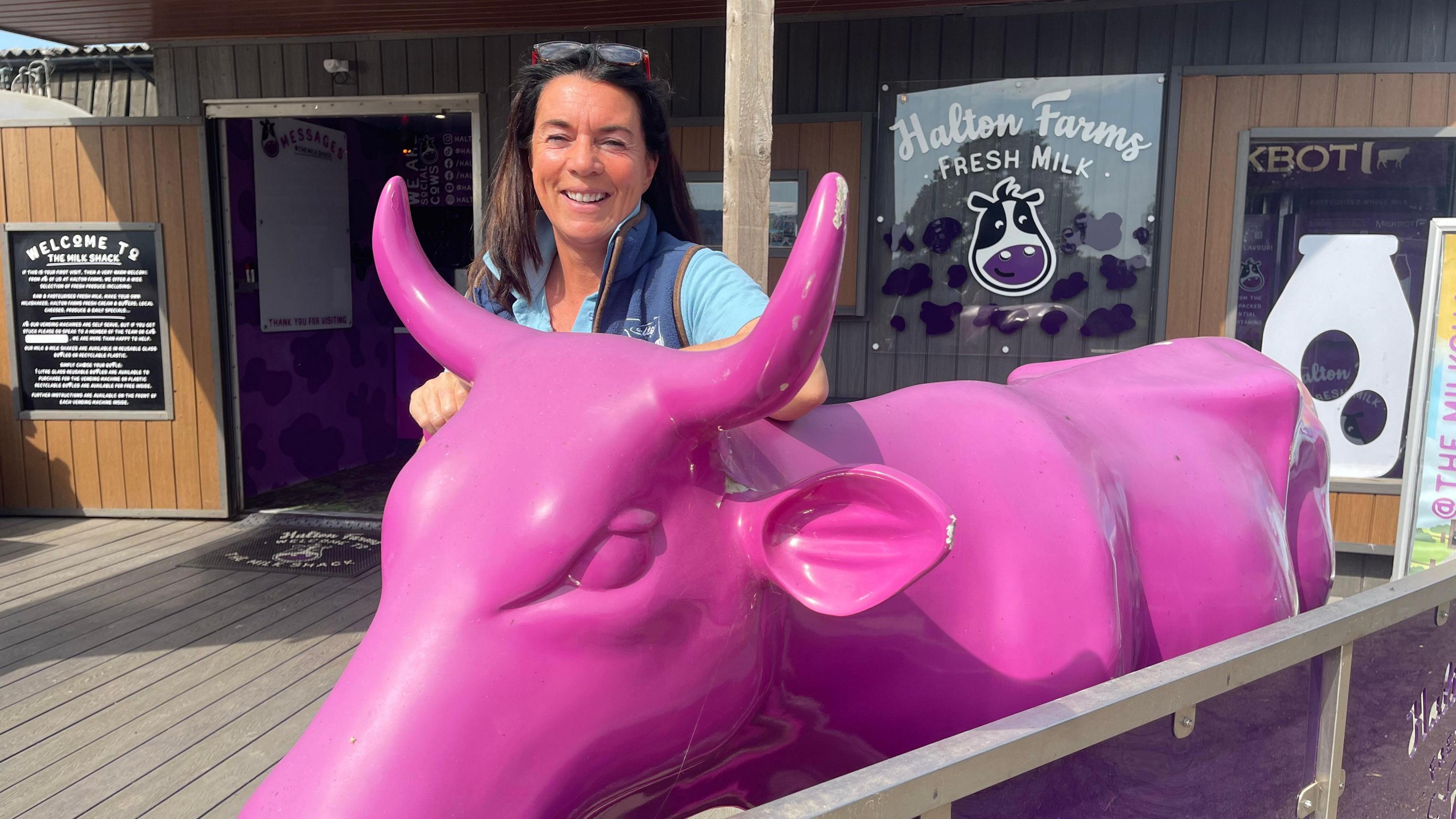 A cheerful female farmer stands behind a purple cow statue. Behind them both is building with an open door and the words ‘Halton Farms Fresh Milk’ printed on the window