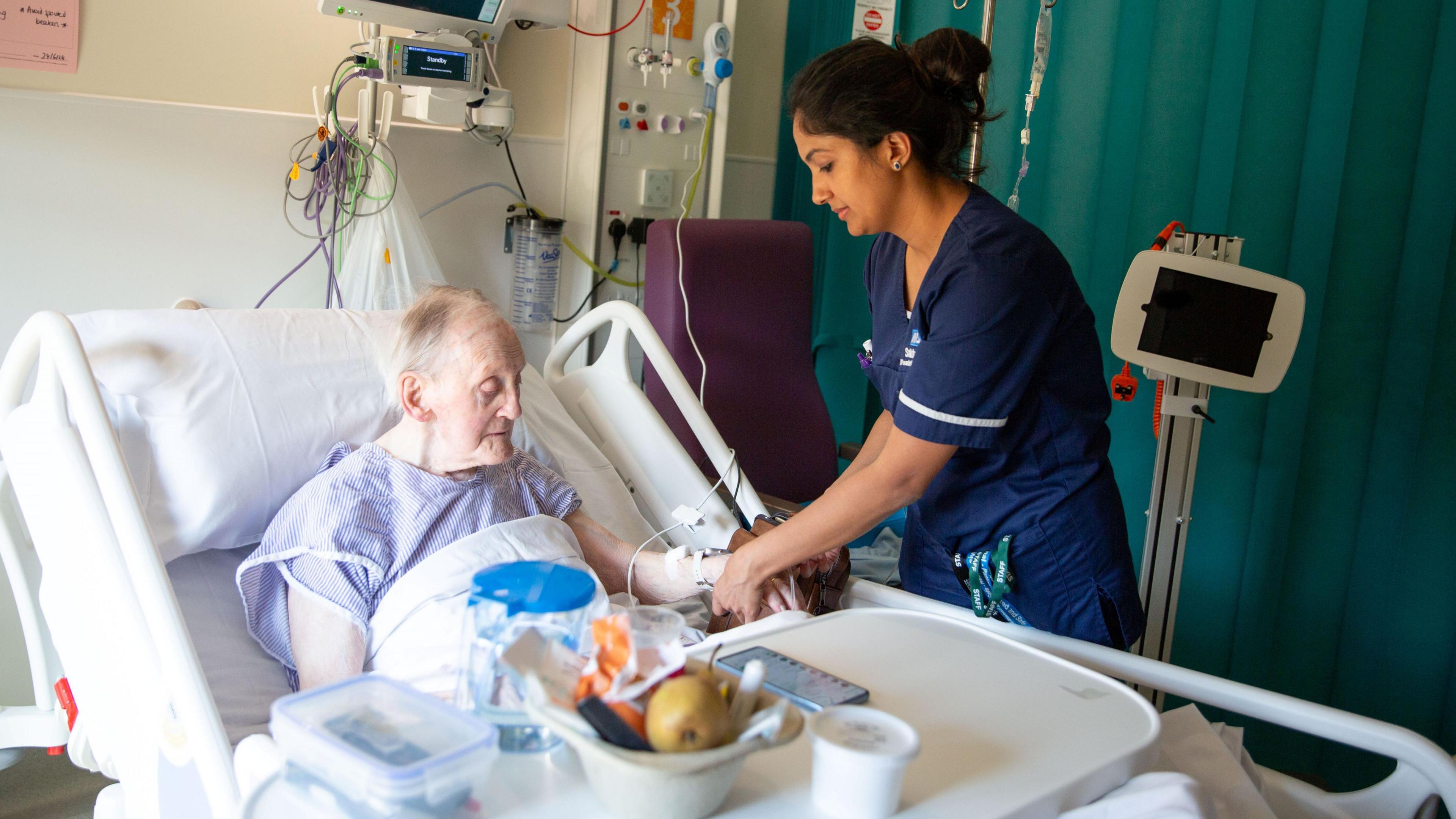 Elderly hospital patient in a bed being checked over by a nurse