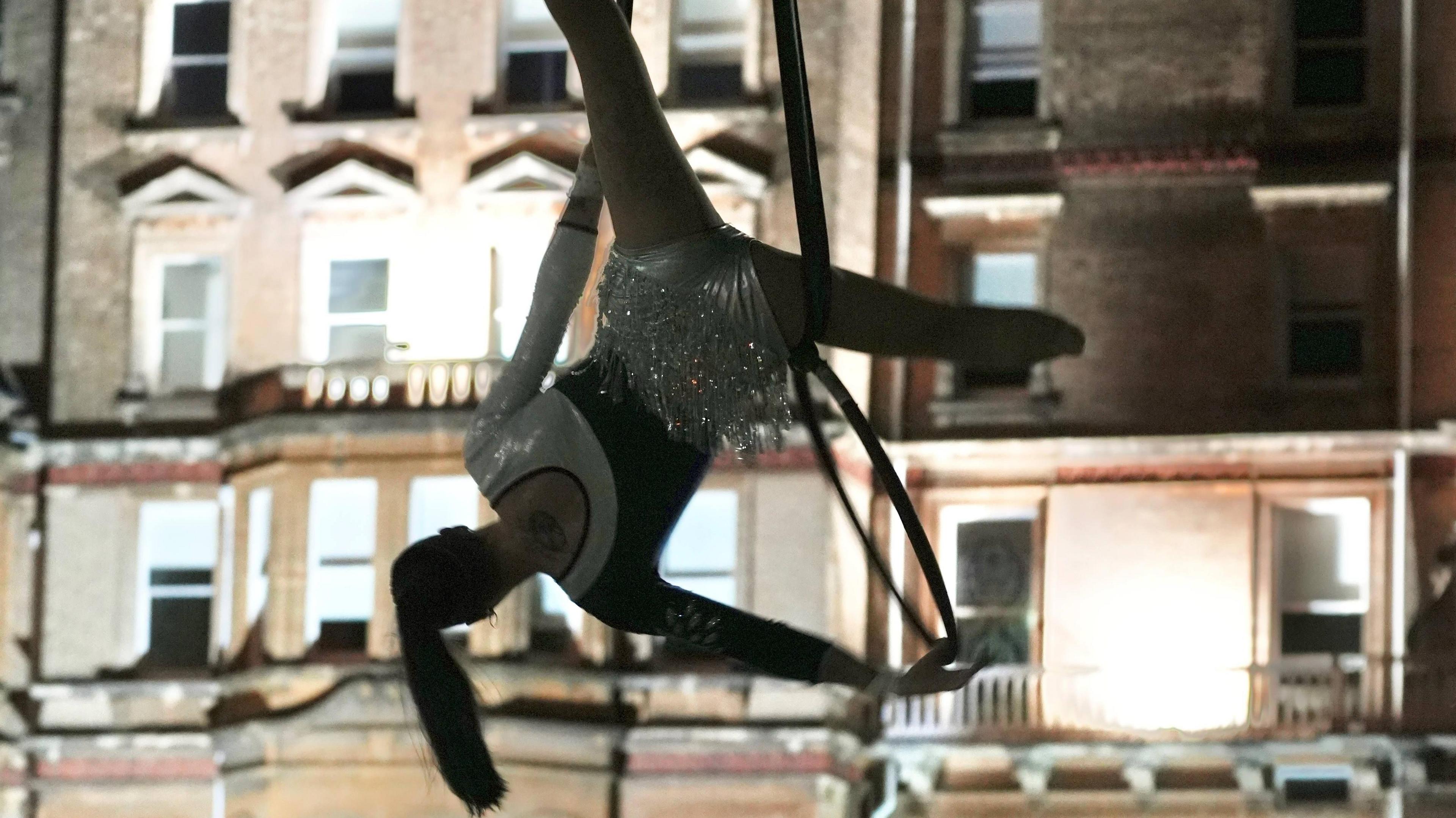 An acrobat hanging from a wire in the evening outside a building. 