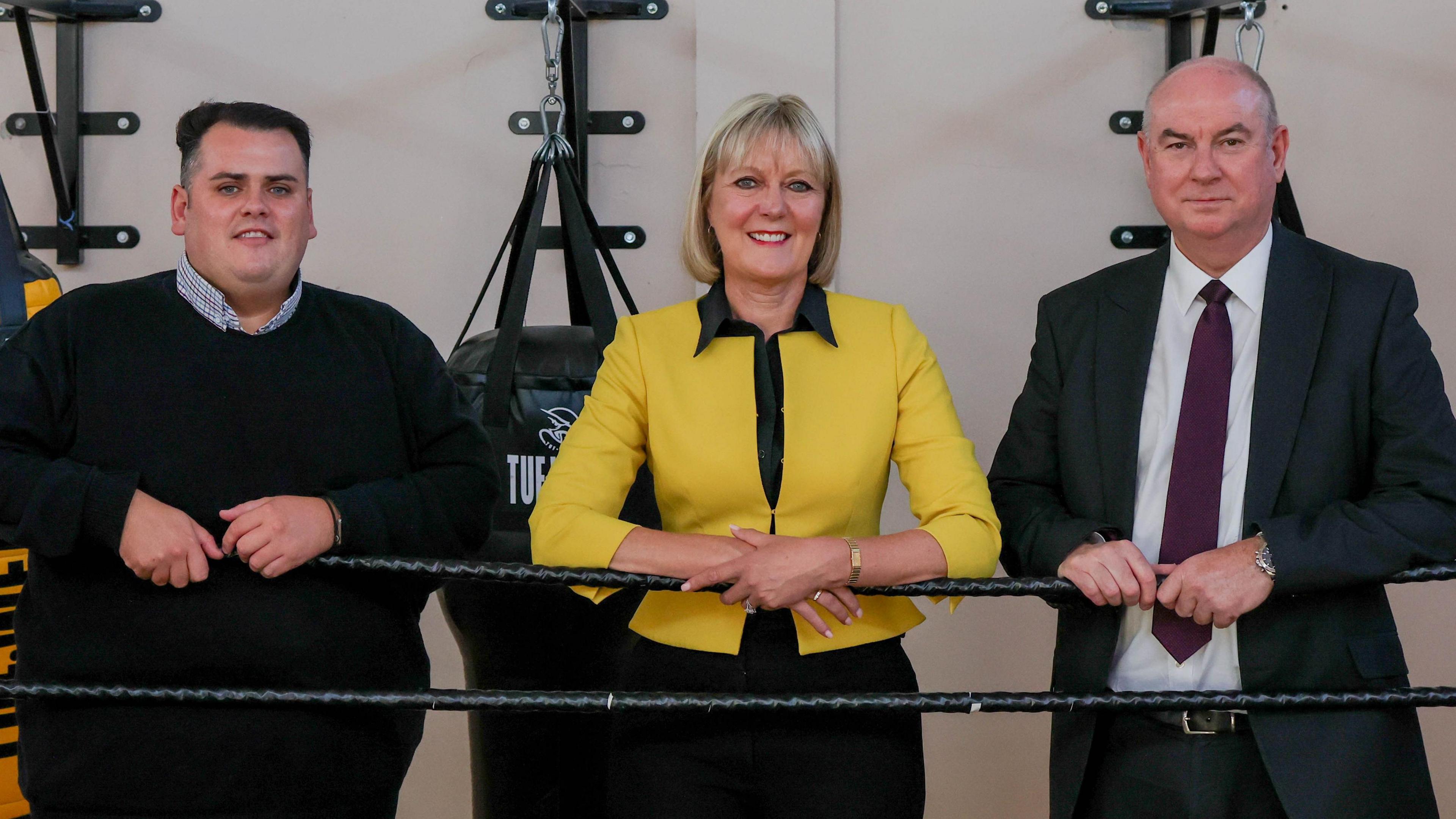 Frome left to right Cllr Jonathan Dulston in a black sweater, Joy Allen PCC in a black shirt and yellow jacket, and Graham Hall Deputy PCC in a black suit and red tie.
They are on the boxing ring at N Gage in Darlington, leaning on the ropes. 
There are boxing punch bags behind them. 