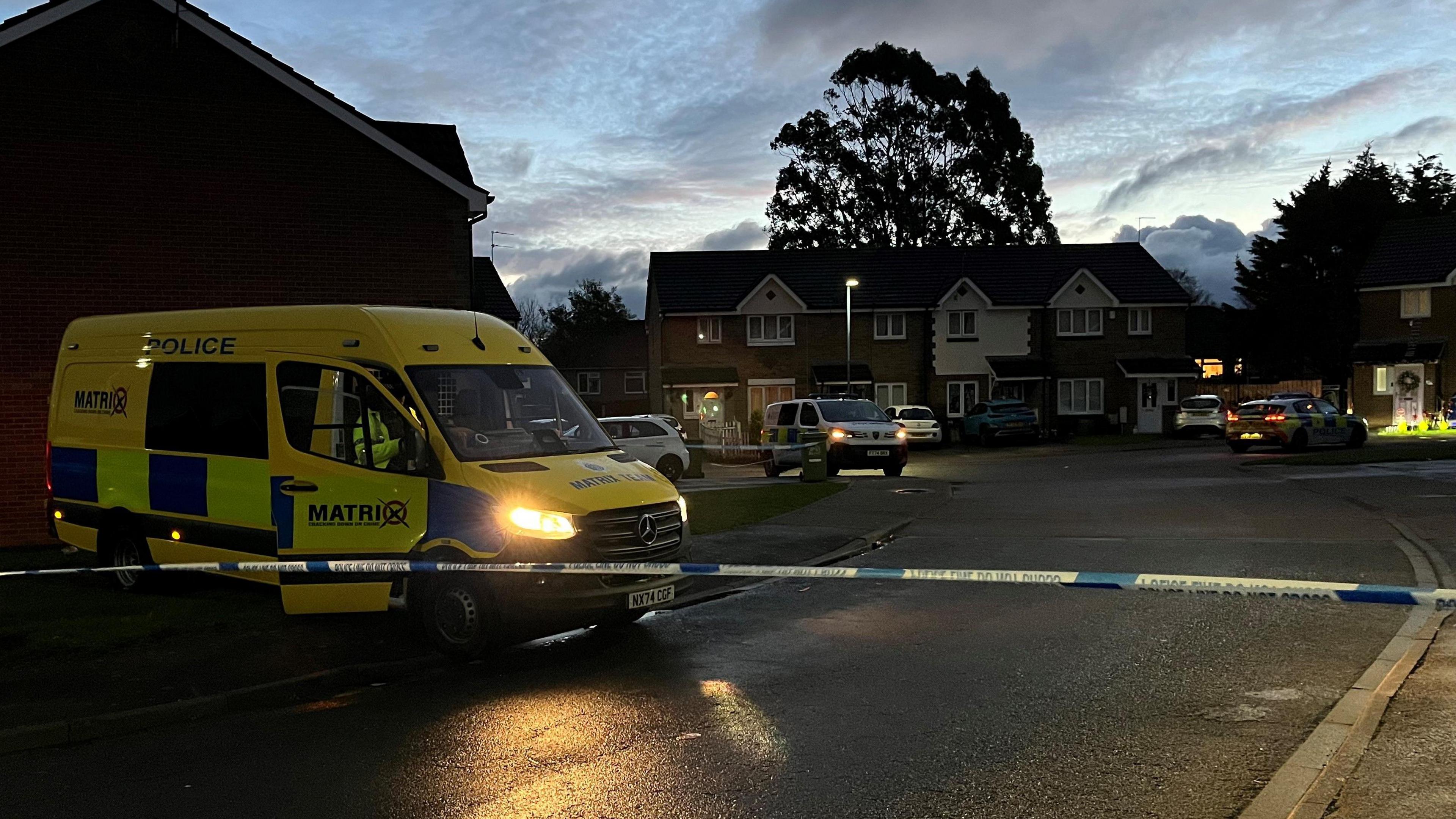 A yellow and blue police van parked behind blue and white police tape with a row of two-storey hoses beyond.