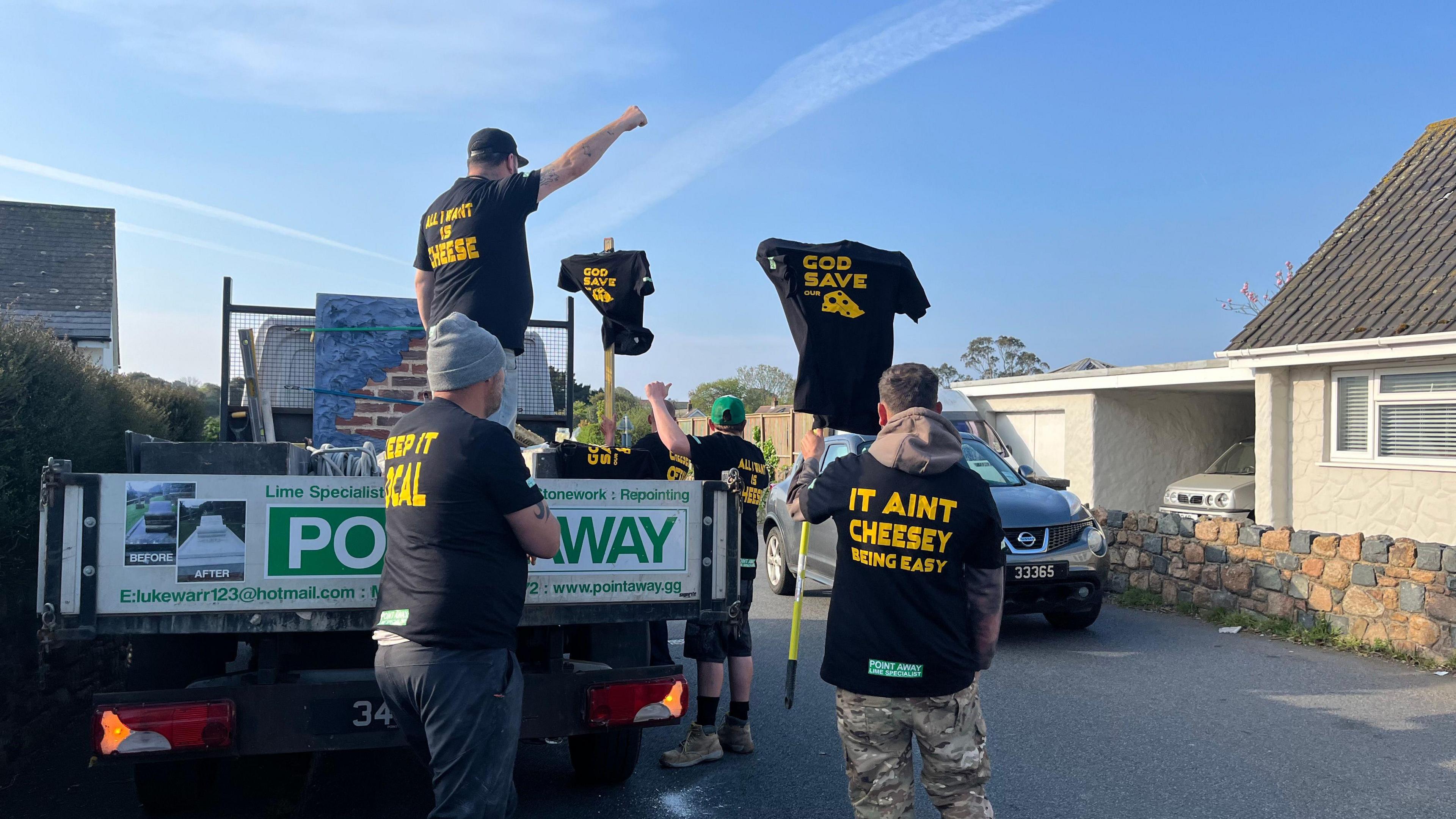 Five mean, wearing black t-shirt with yellow writing on them, next to a van parked on a road. Opposite them is a Nissan car and a house. They are all facing away from the camera. Two of the men are holding up t-shirts on poles with slogans on. Some of the slogans read "it ain't cheesey being easy" and "God save the cheese"