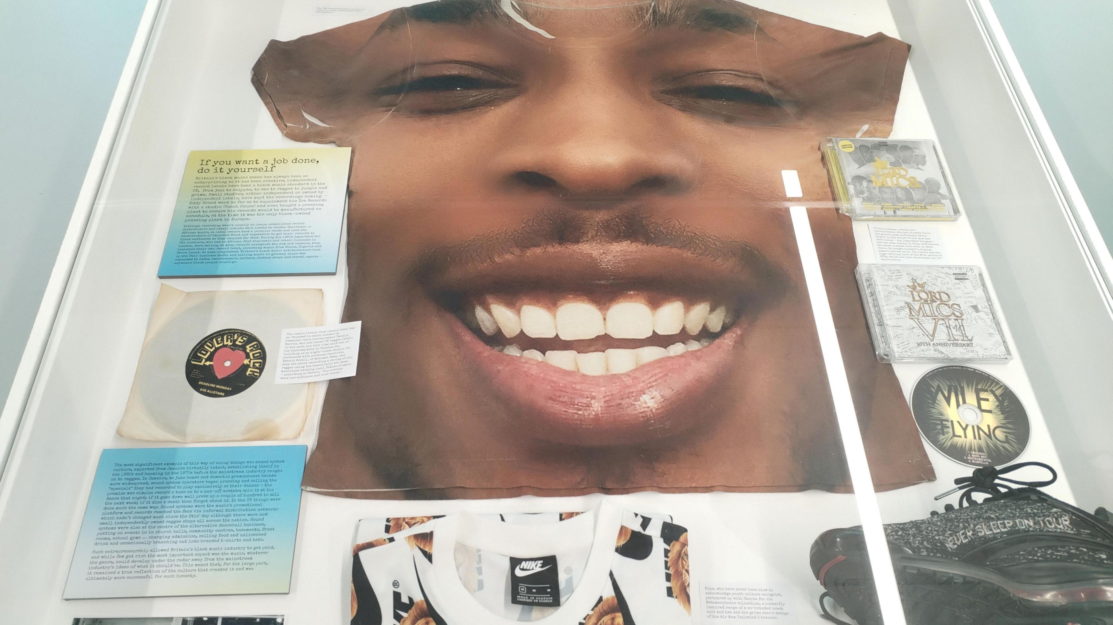 A photo of one of the exhibits with a large T-shirt with a man smiling on it, a vinyl, some CDs and some black trainers. 