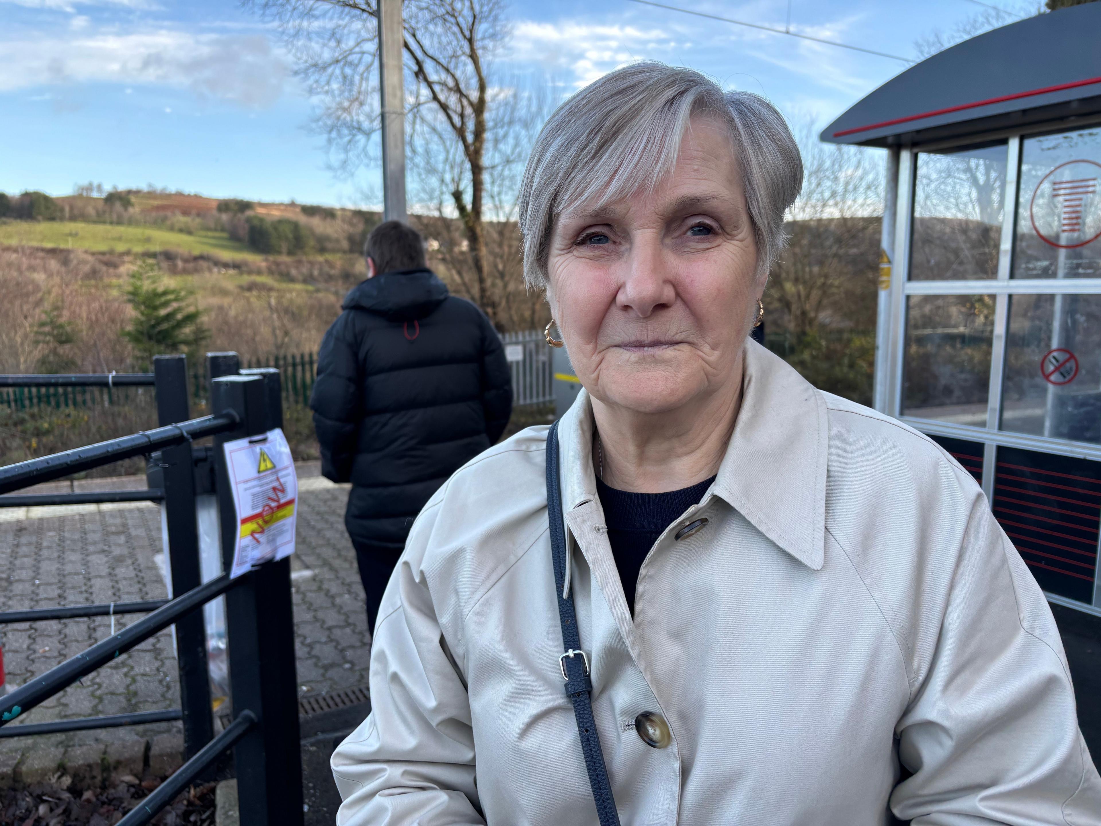 Menyw gwallt llwyd mewn côt wen yn sefyll ar bwys gorsaf drenau Pontlotyn