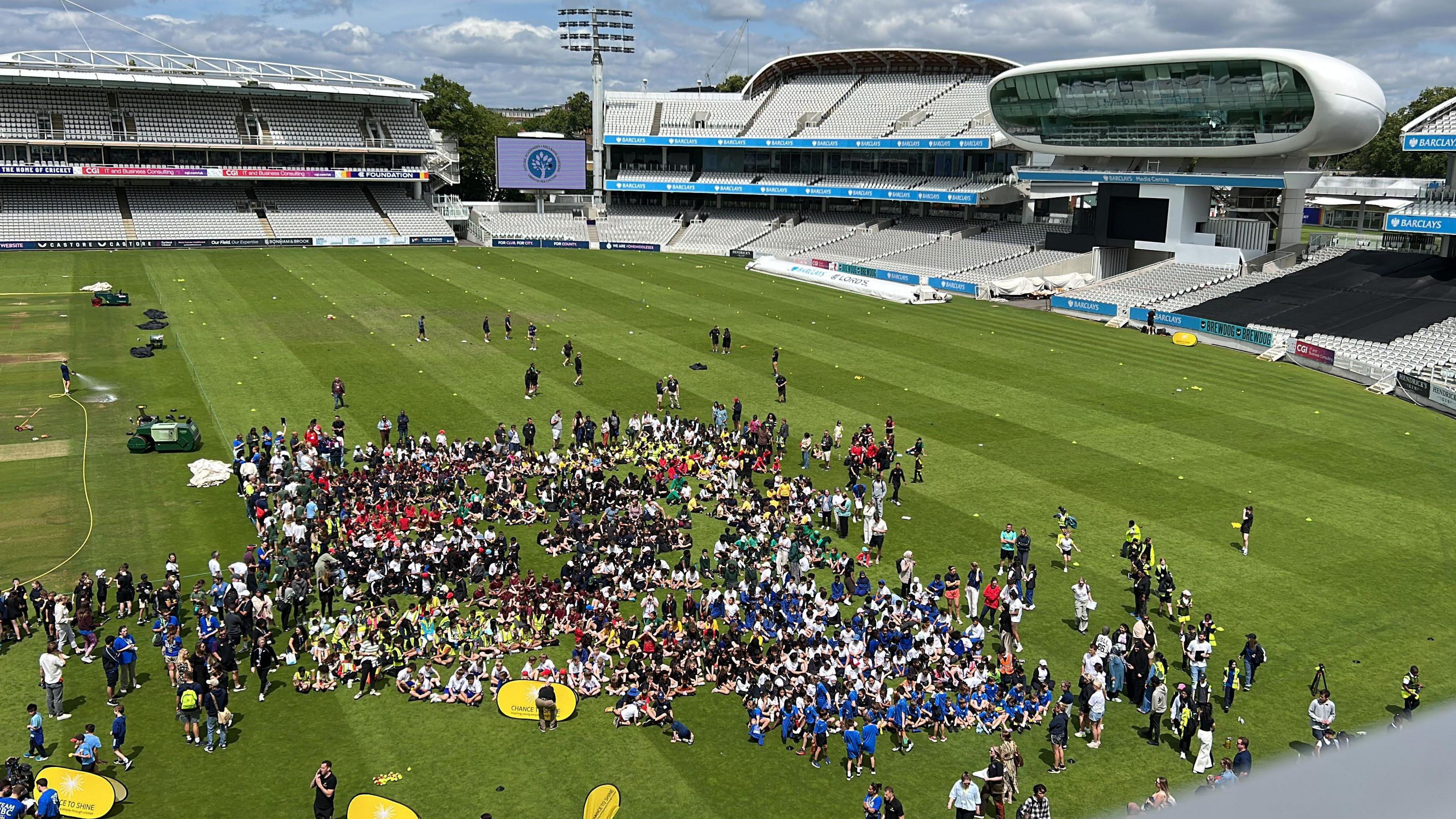 A large group of young people stand and sit on the grass at Lord's cricket ground. The stands are in the background.