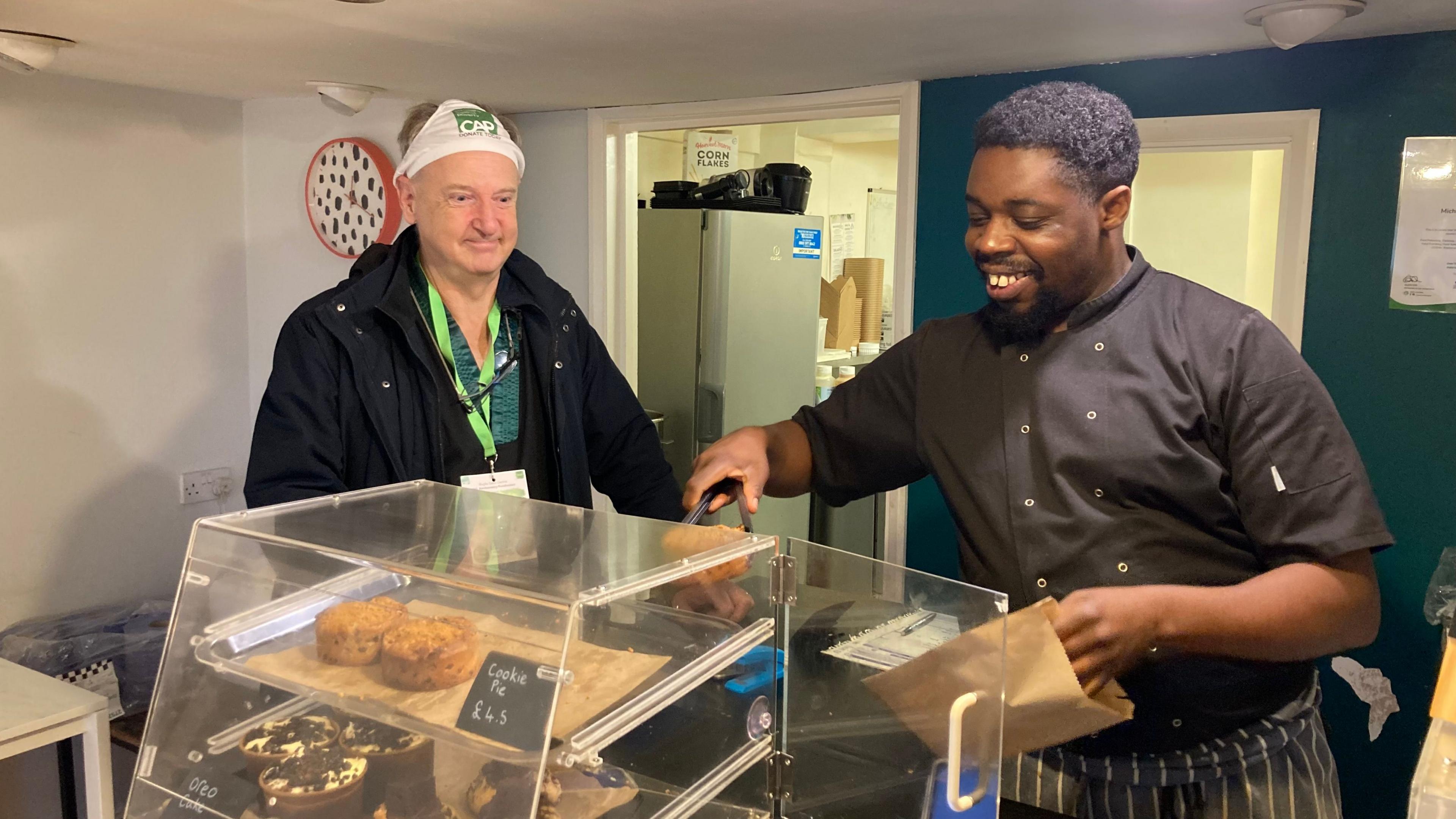 Michael Okundaye with short dark hair wearing dark grey shirt and taking a cake from a clear display cabinet, watched by Mark Robson with white pants on his head in a bakery.