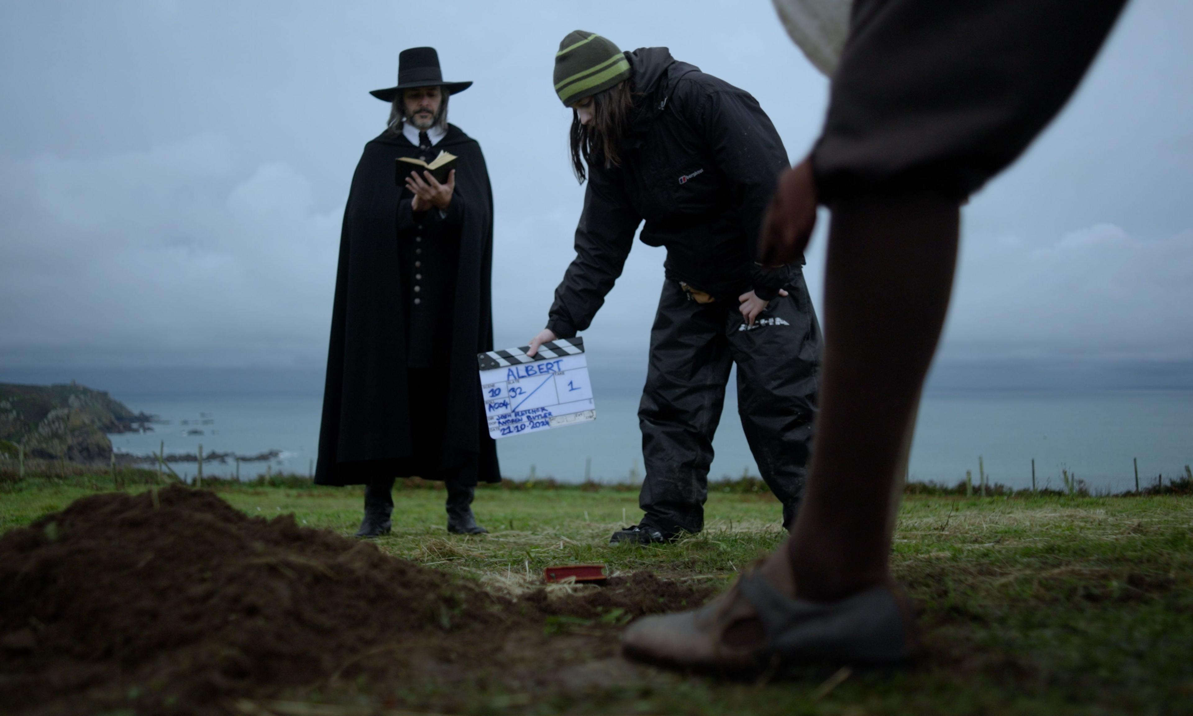 An actor standing in a field on a cliff dressed in a historical black puritan's outfit and hat, holding up a small book. A member of the film crew holds up a clapper board saying: Albert. Another actor's shoe and leg are in view.