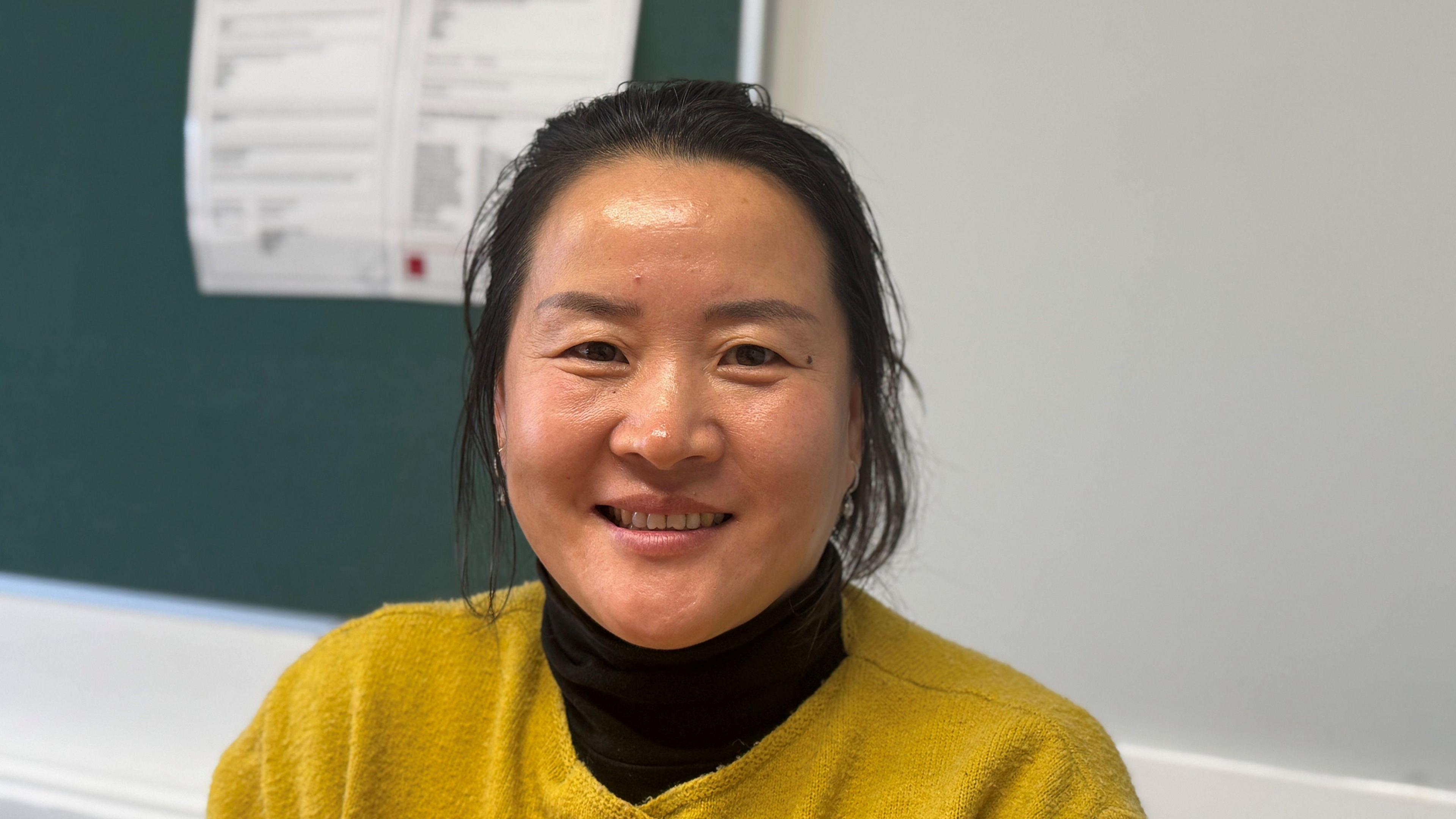 A woman with black hair tied back is smiling at the camera. She has a yellow jumper on, with a black turtleneck underneath. There is a dark green noticeboard behind her.