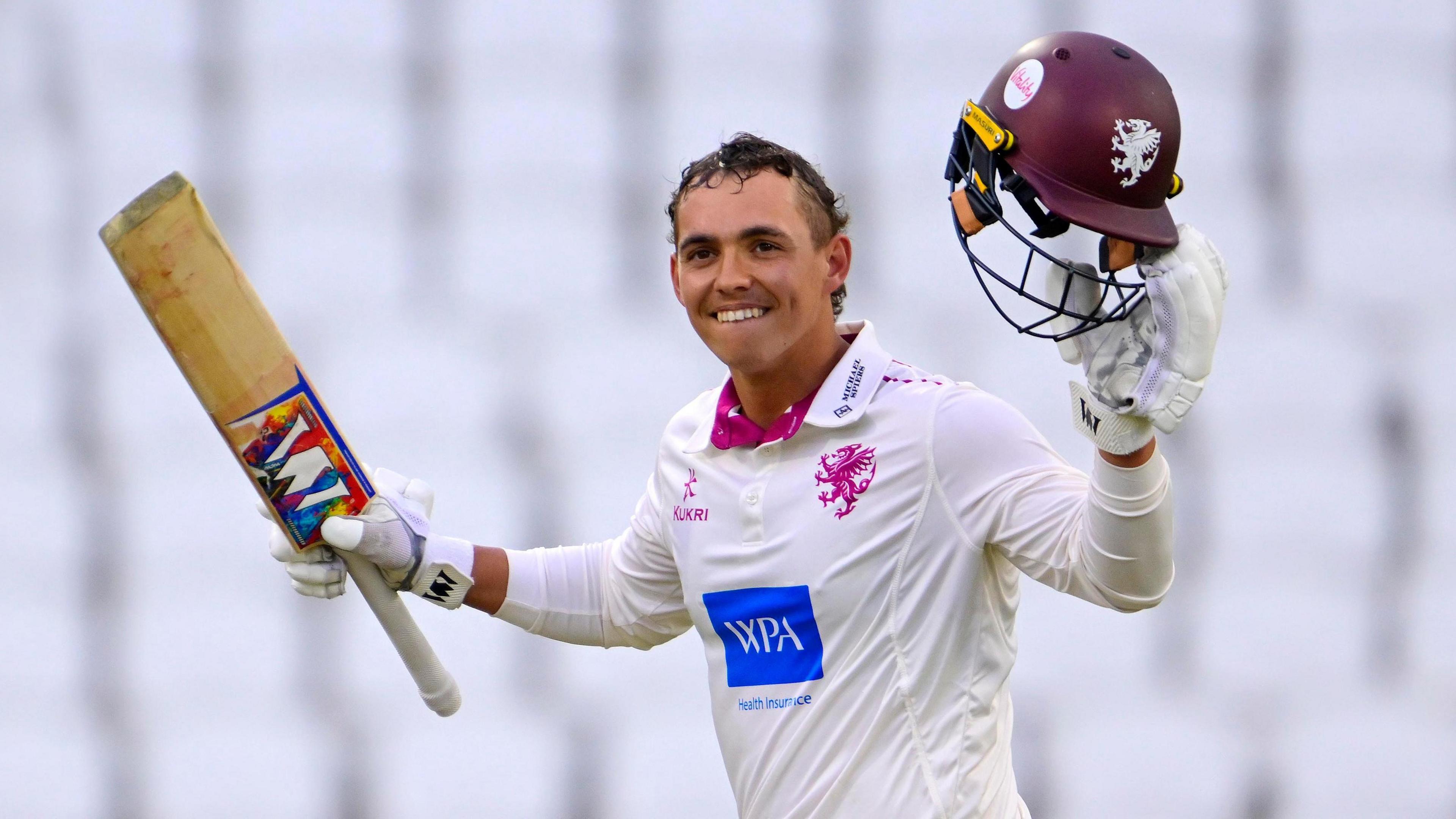 Josh Thomas holds his bat in the air in his right hand and his helmet in his left as he smiles after scoring a century against Yorkshire