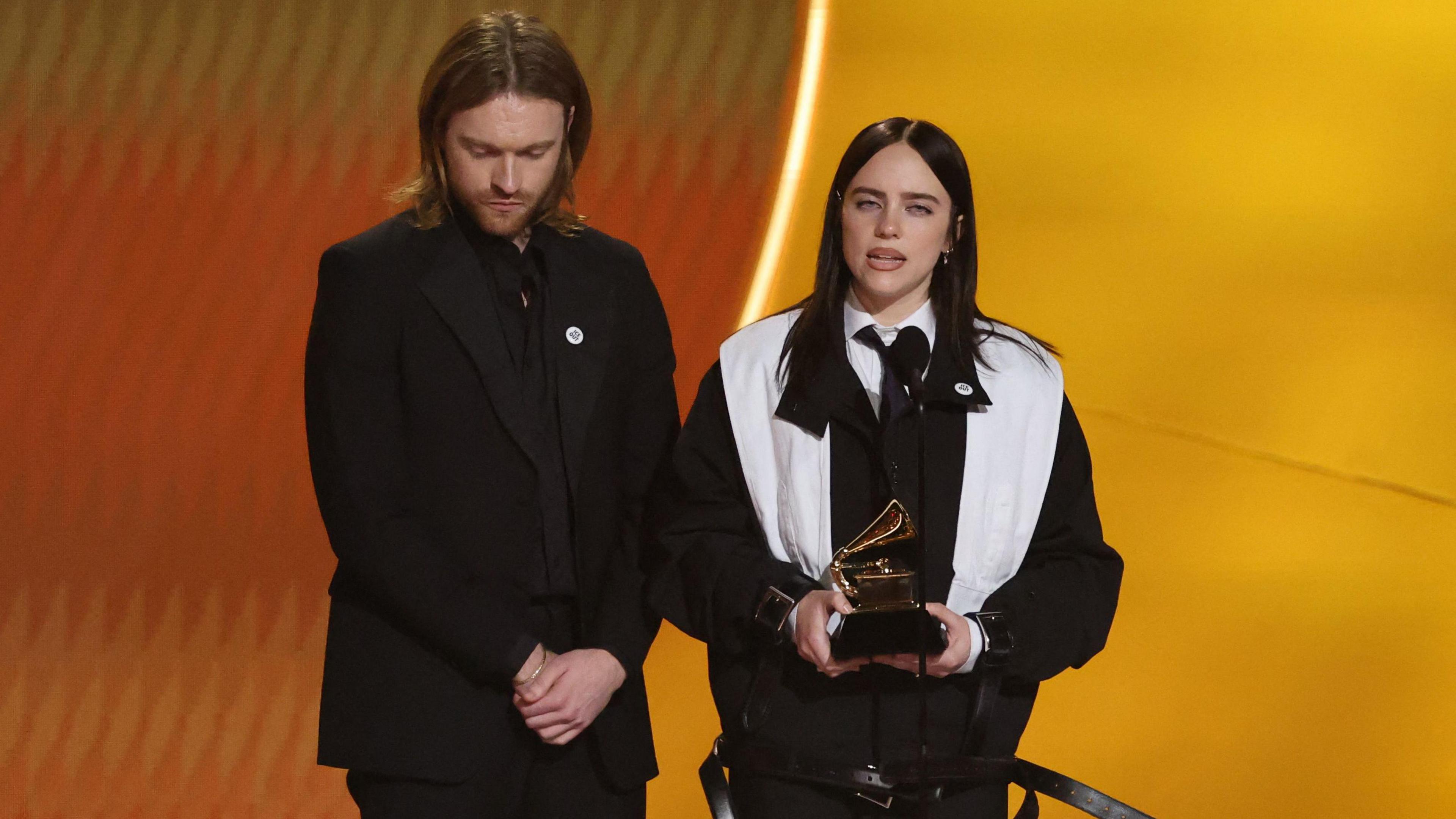 Billie Eilish is standing on stage holding a grammy as she accepts the award for Song of the Year with her brother and producer Finneas standing on the left 